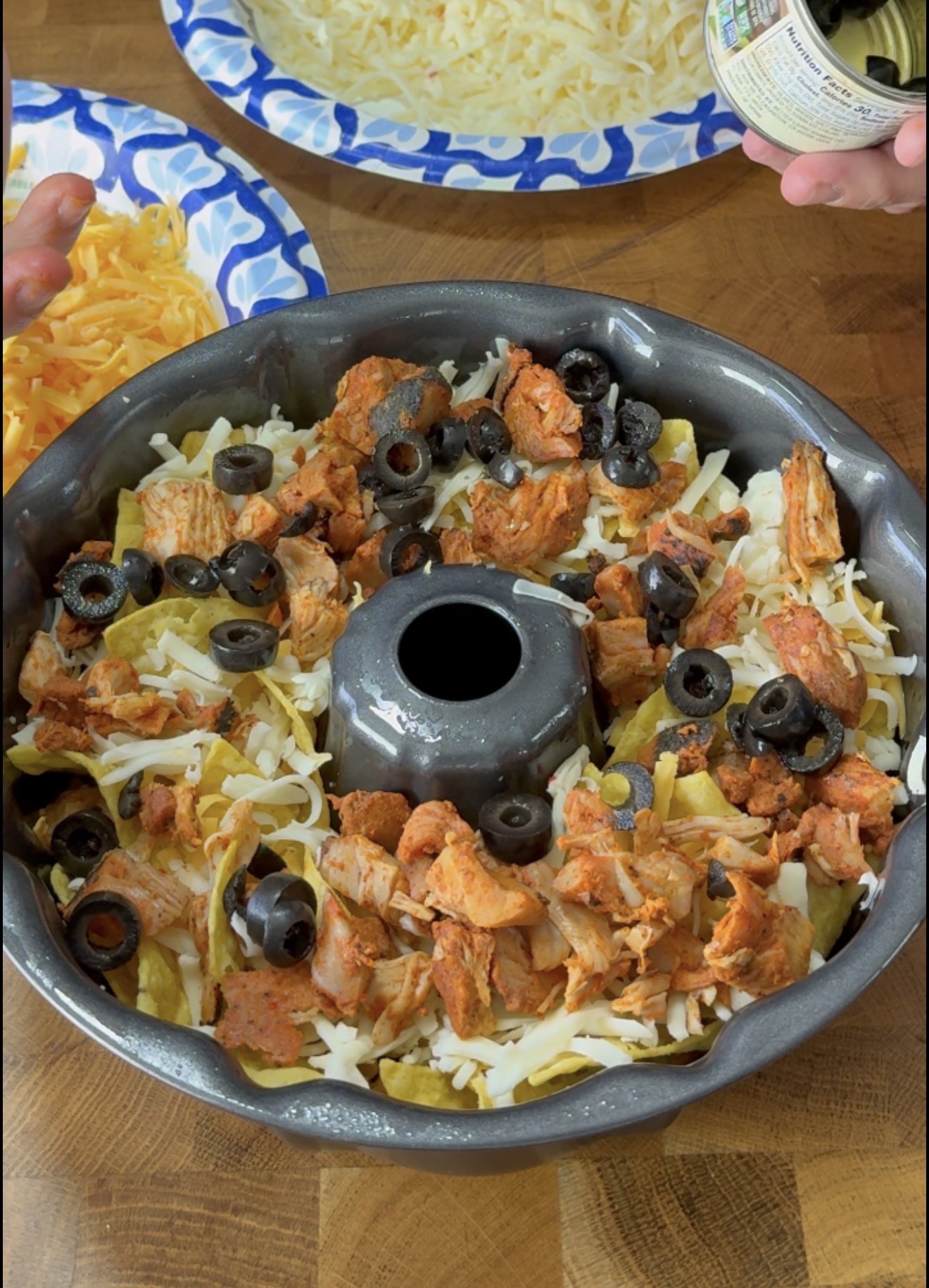 A bundt pan filled with layers of shredded cheese, seasoned chicken, green chilies, and sliced black olives sits on a wooden counter. Paper plates with more cheese are nearby, and a hand holds an open can at the edge.