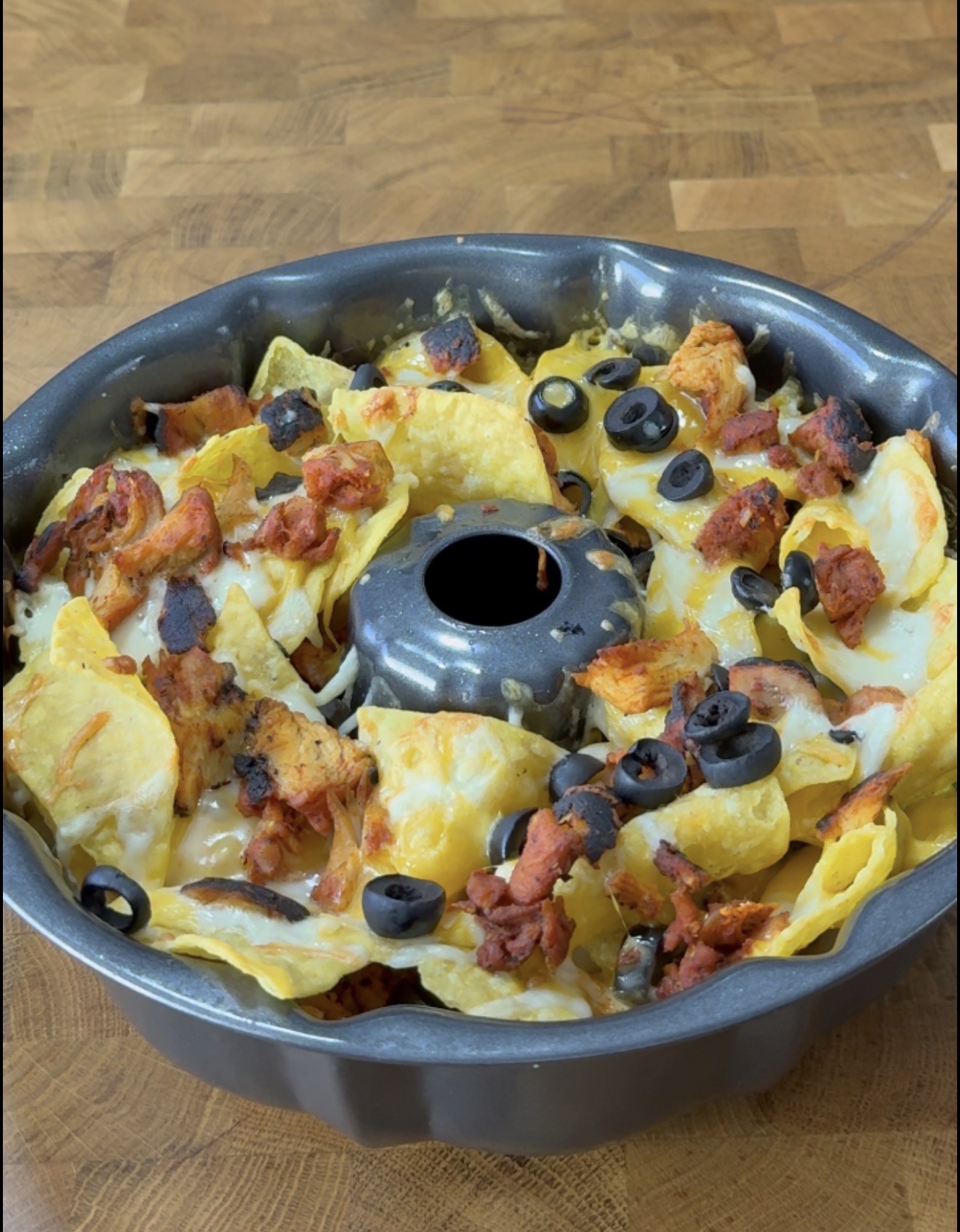 A bundt pan filled with nachos, topped with melted cheese, grilled chicken pieces, chopped tomatoes, and sliced black olives, sits on a wooden surface.