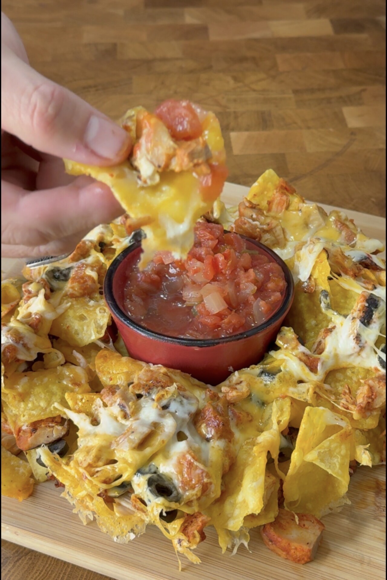 A hand dips a cheesy nacho chip topped with chicken into a bowl of salsa, surrounded by a platter of loaded nachos on a wooden surface.