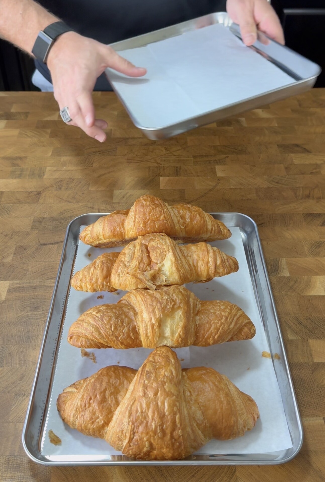 A person holds an empty baking tray above a wooden counter. In front, four golden-brown croissants are arranged on a parchment-lined tray.