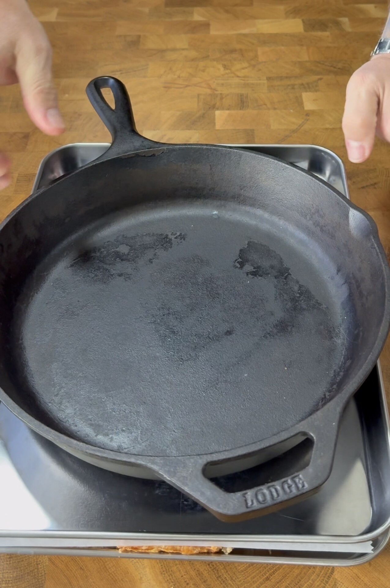A person places a black cast iron skillet labeled Lodge onto a shiny metal tray set on a wooden countertop.