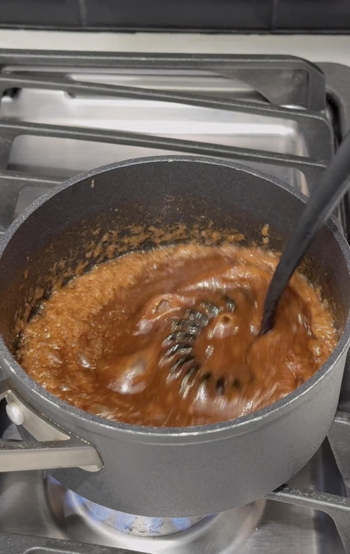 A black pot on a stove with a brown, bubbling mixture being stirred by a black utensil.