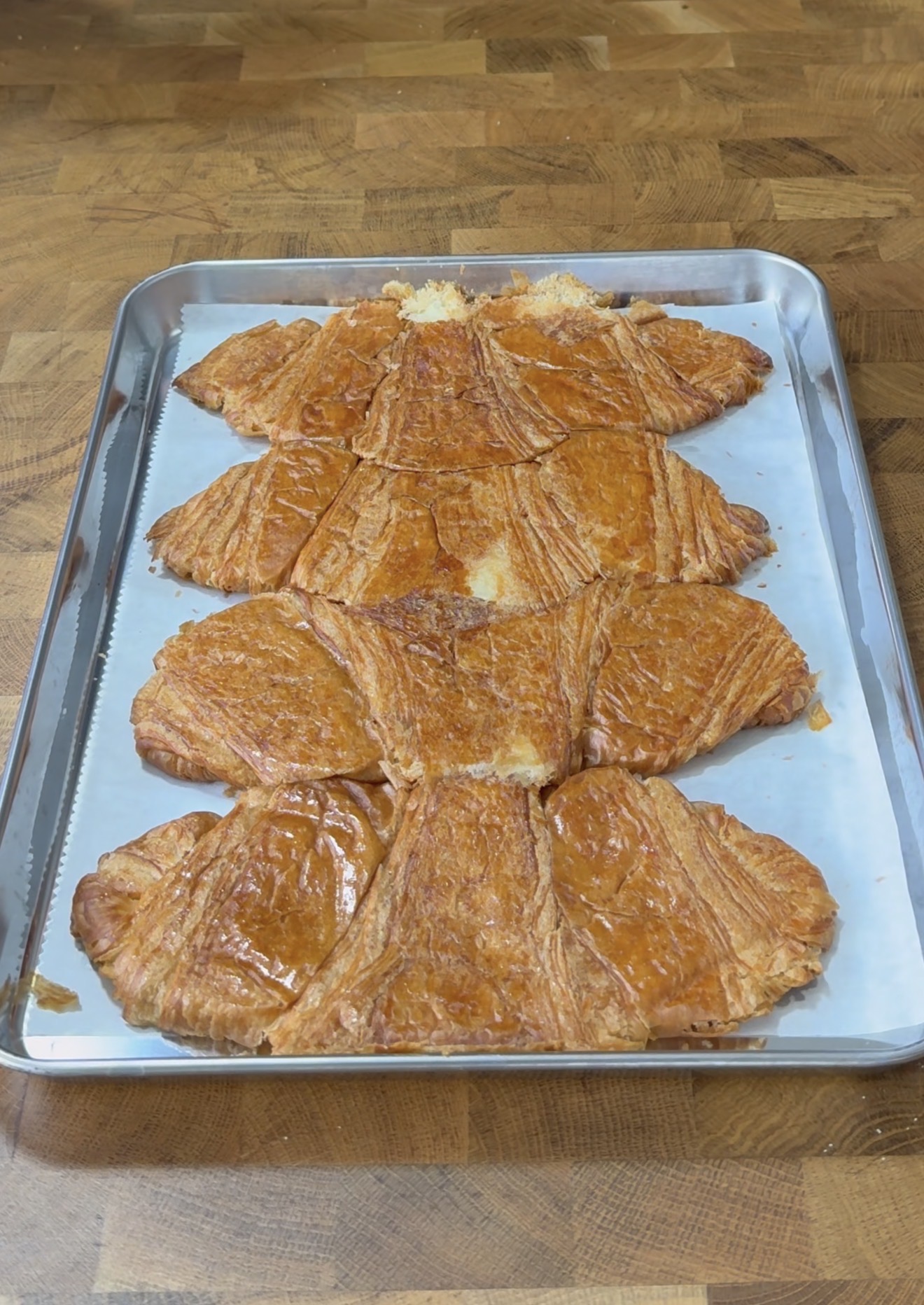 A large baked pastry shaped like a teddy bear, with a shiny golden-brown crust, sits on parchment paper atop a metal baking tray on a wooden surface.