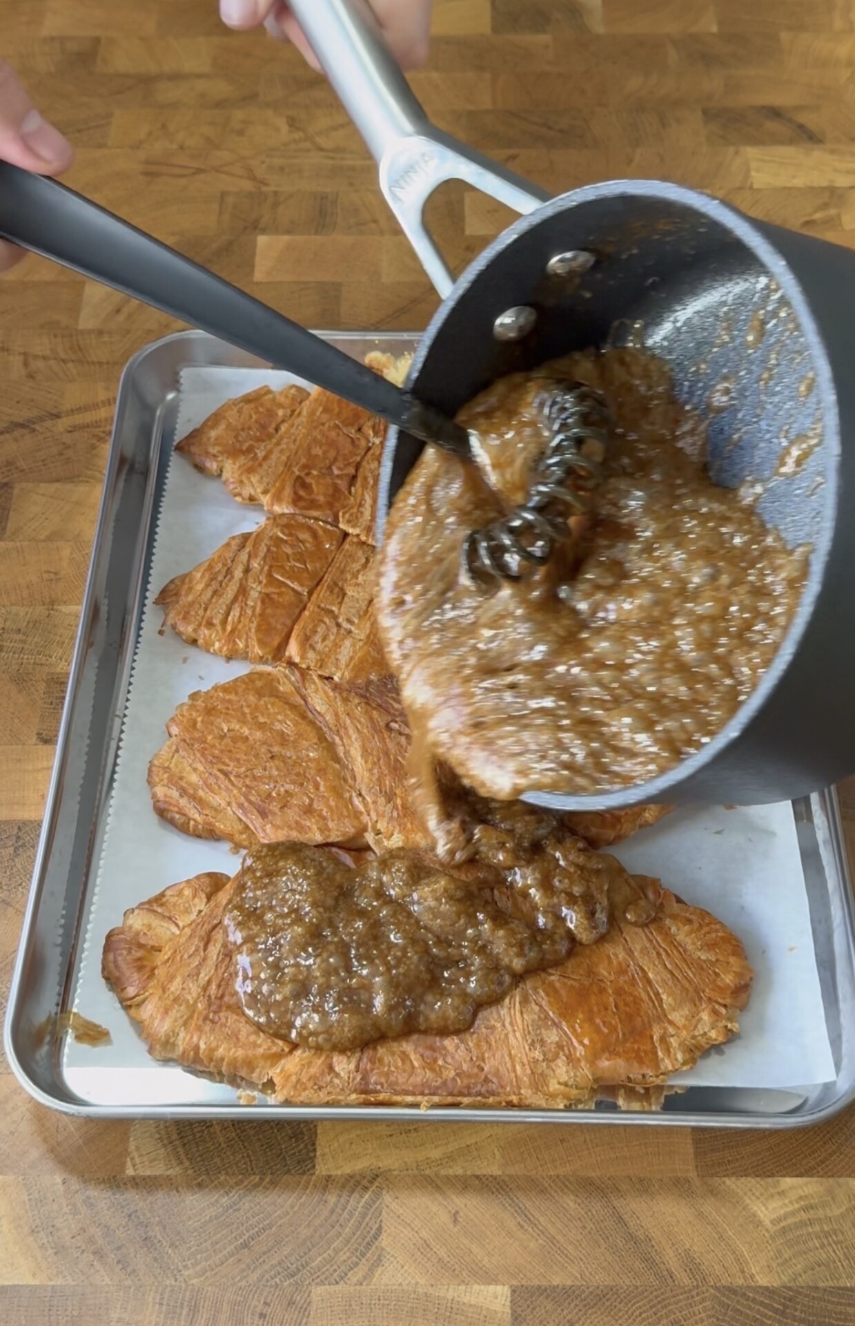A saucepan pours a thick, brown mixture over sliced croissants on a parchment-lined baking sheet, set on a wooden surface.