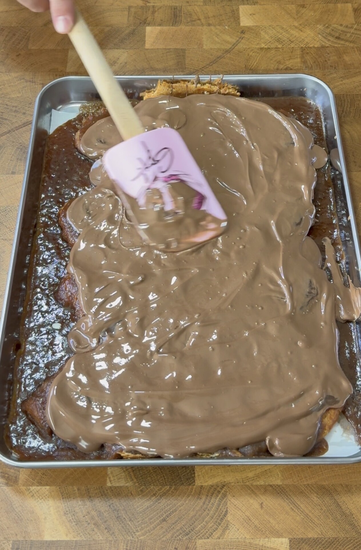 A hand spreads melted chocolate over a large rectangular sheet cake using a pink spatula on a metal tray, atop a wooden surface.
