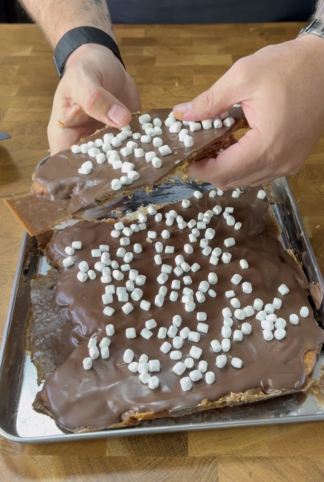 A person breaks apart a sheet of chocolate-covered dessert topped with mini marshmallows on a metal tray, on a wooden surface.