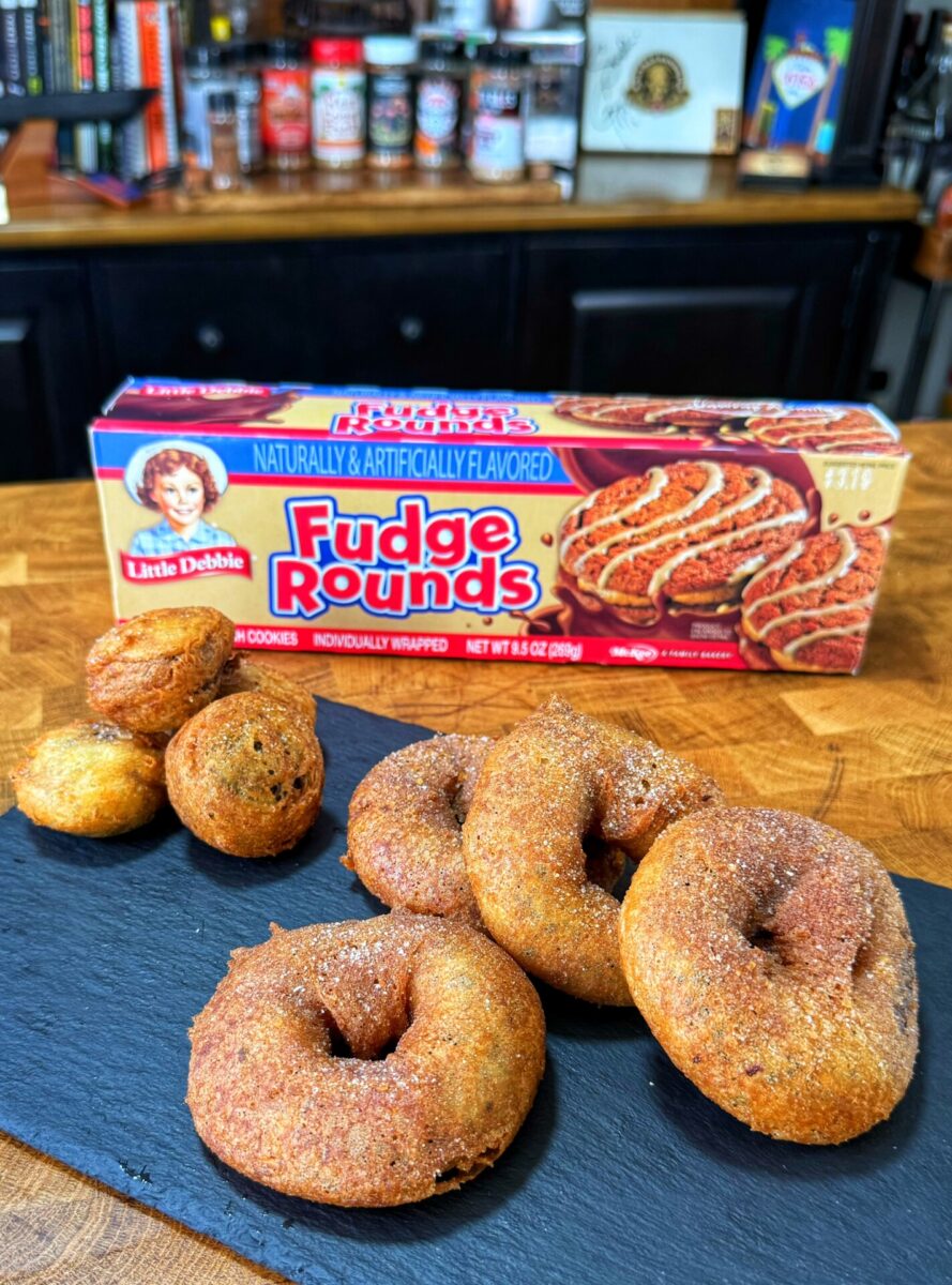 A slate tray with homemade donuts and donut holes sits in front of a box of Little Debbie Fudge Rounds on a wooden counter, with spice jars and kitchen items in the background.