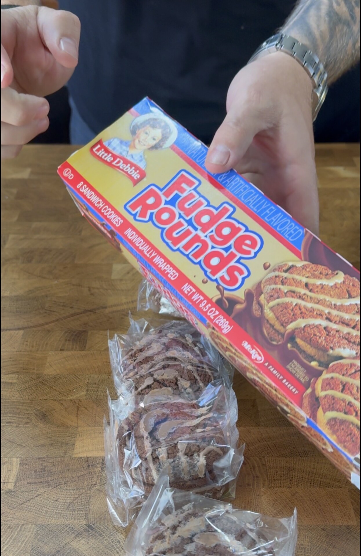 A person opens a Little Debbie Fudge Rounds box on a wooden table, with several individually wrapped Fudge Rounds snacks visible in front of them.