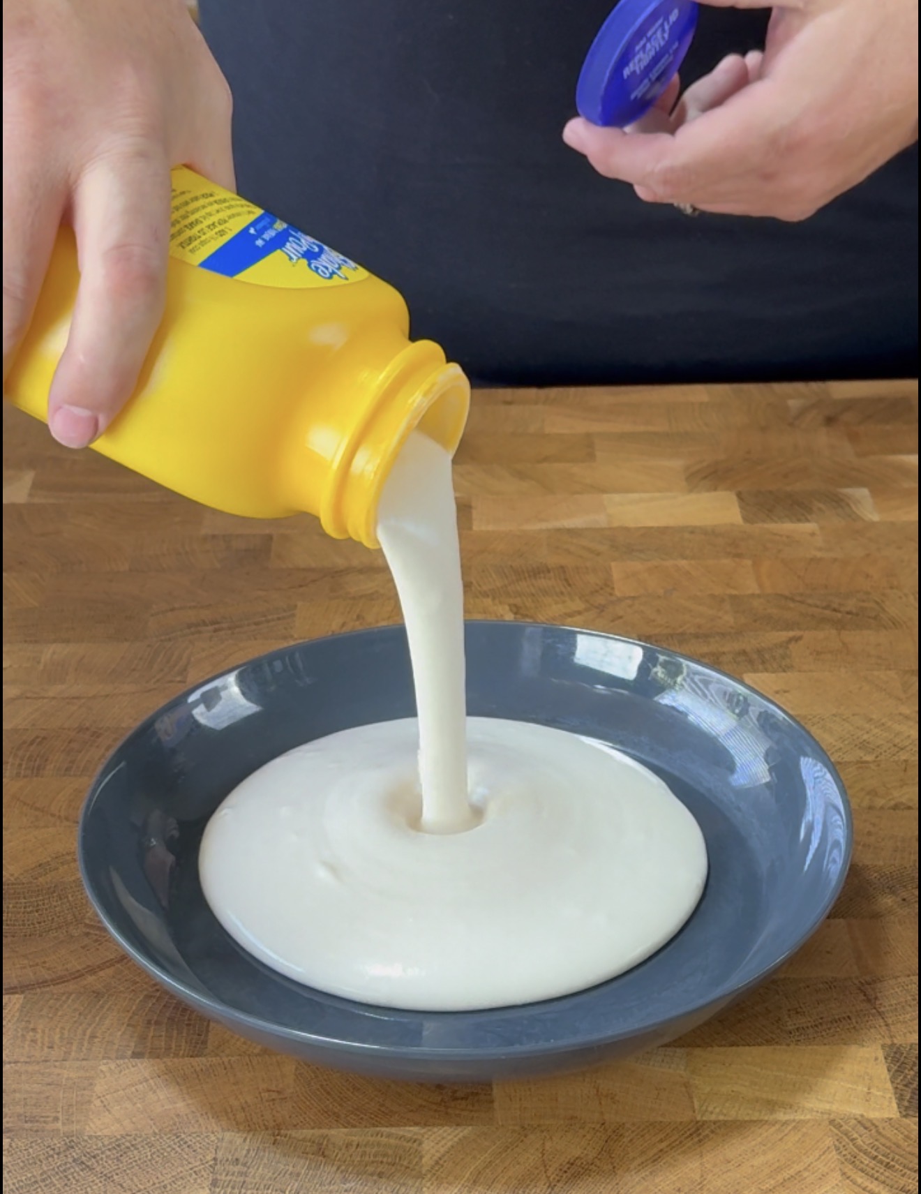A person pours a thick white liquid from a yellow squeeze bottle into a dark gray bowl on a wooden surface.