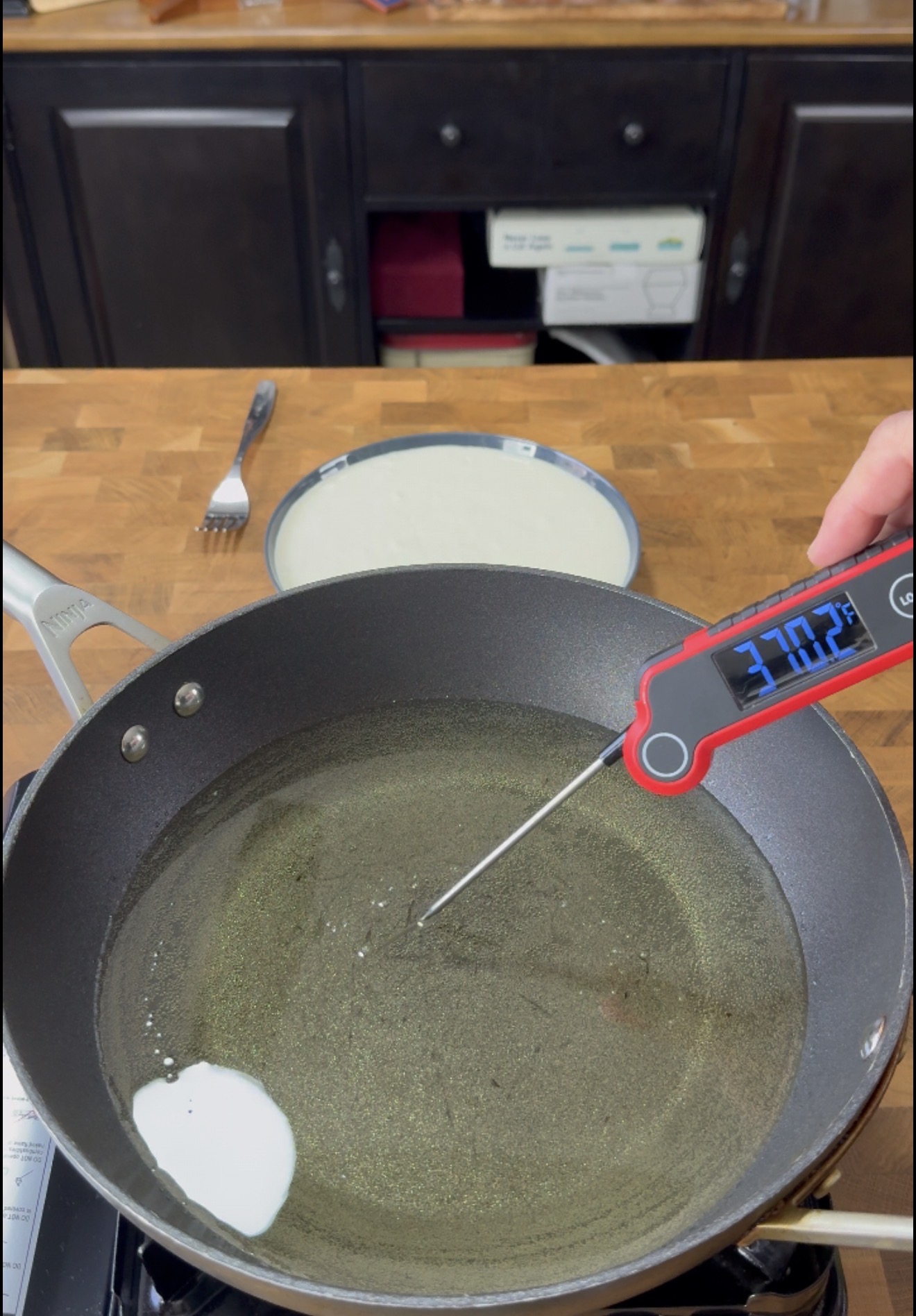 A hand holds a digital thermometer in a frying pan with oil reading 372.7°F. A fork and a plate with batter are on a wooden counter in the background.