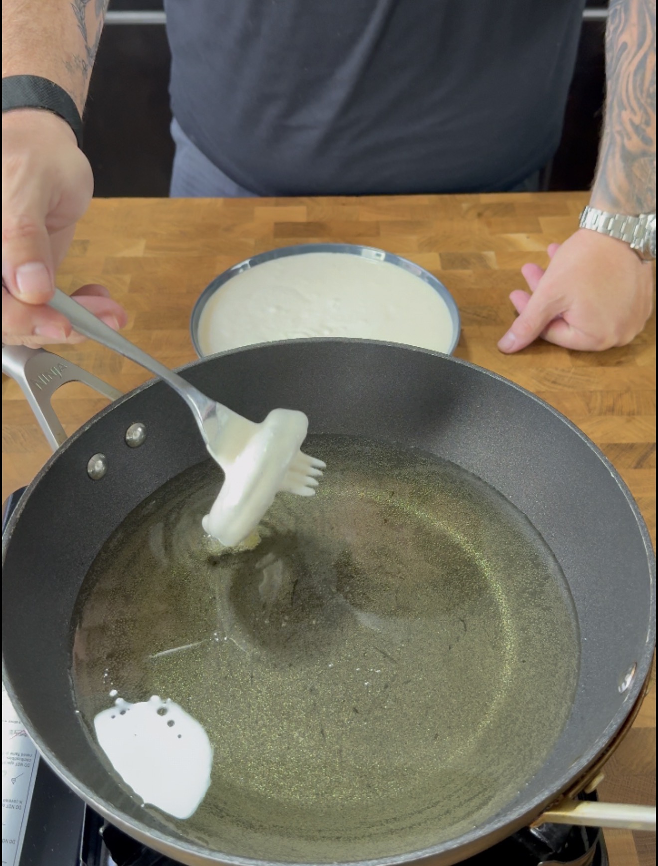 A person holds a fork with batter over a pan of hot oil, preparing to fry. A bowl of batter sits on a wooden countertop.