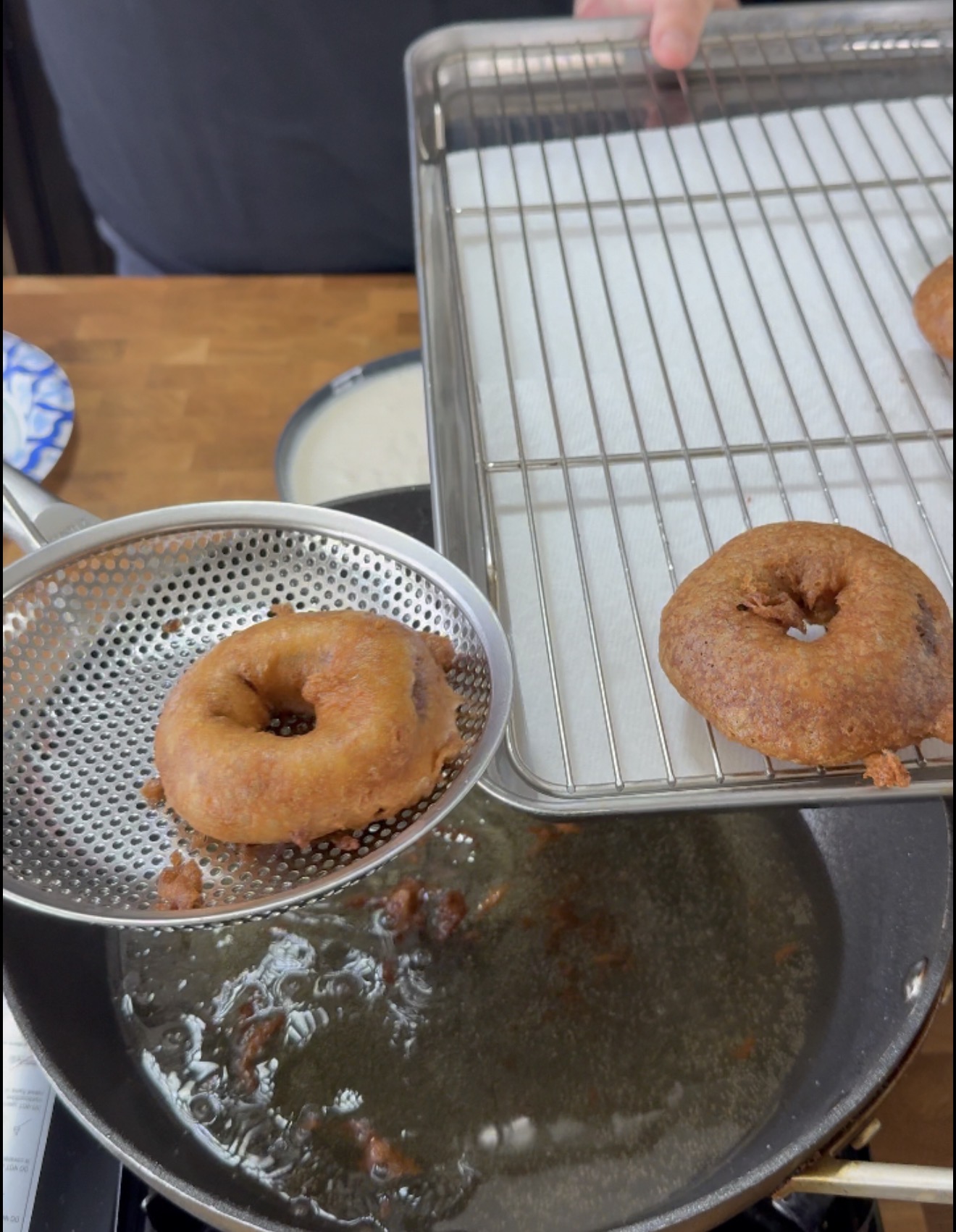 A person transfers a freshly fried donut from a slotted skimmer to a cooling rack lined with paper towels over a pan of hot oil.