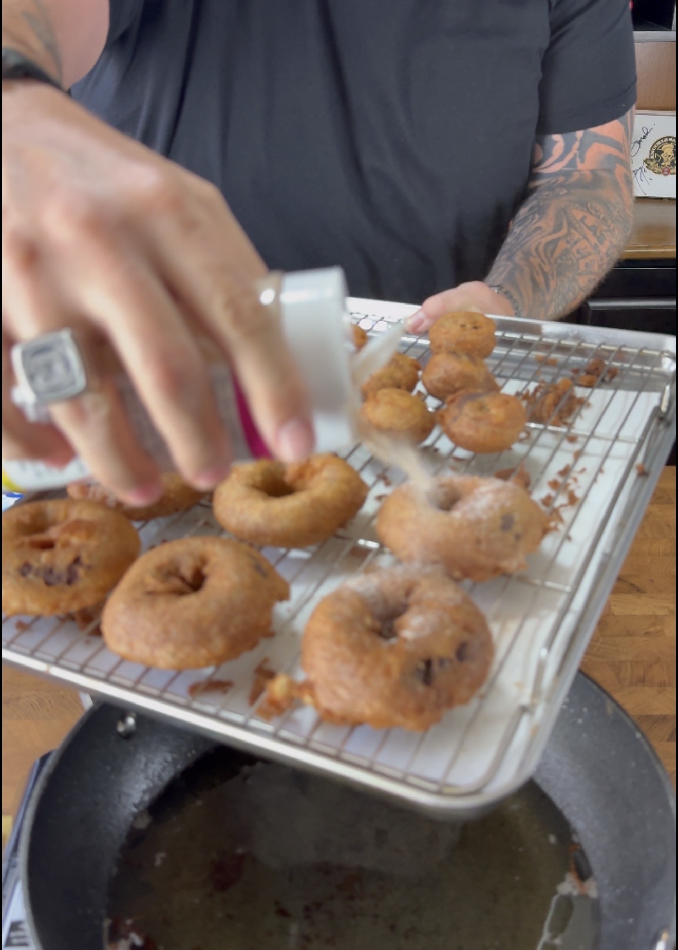 A tattooed person sprinkles sugar onto freshly fried donuts on a wire rack over a baking sheet, with a frying pan below.