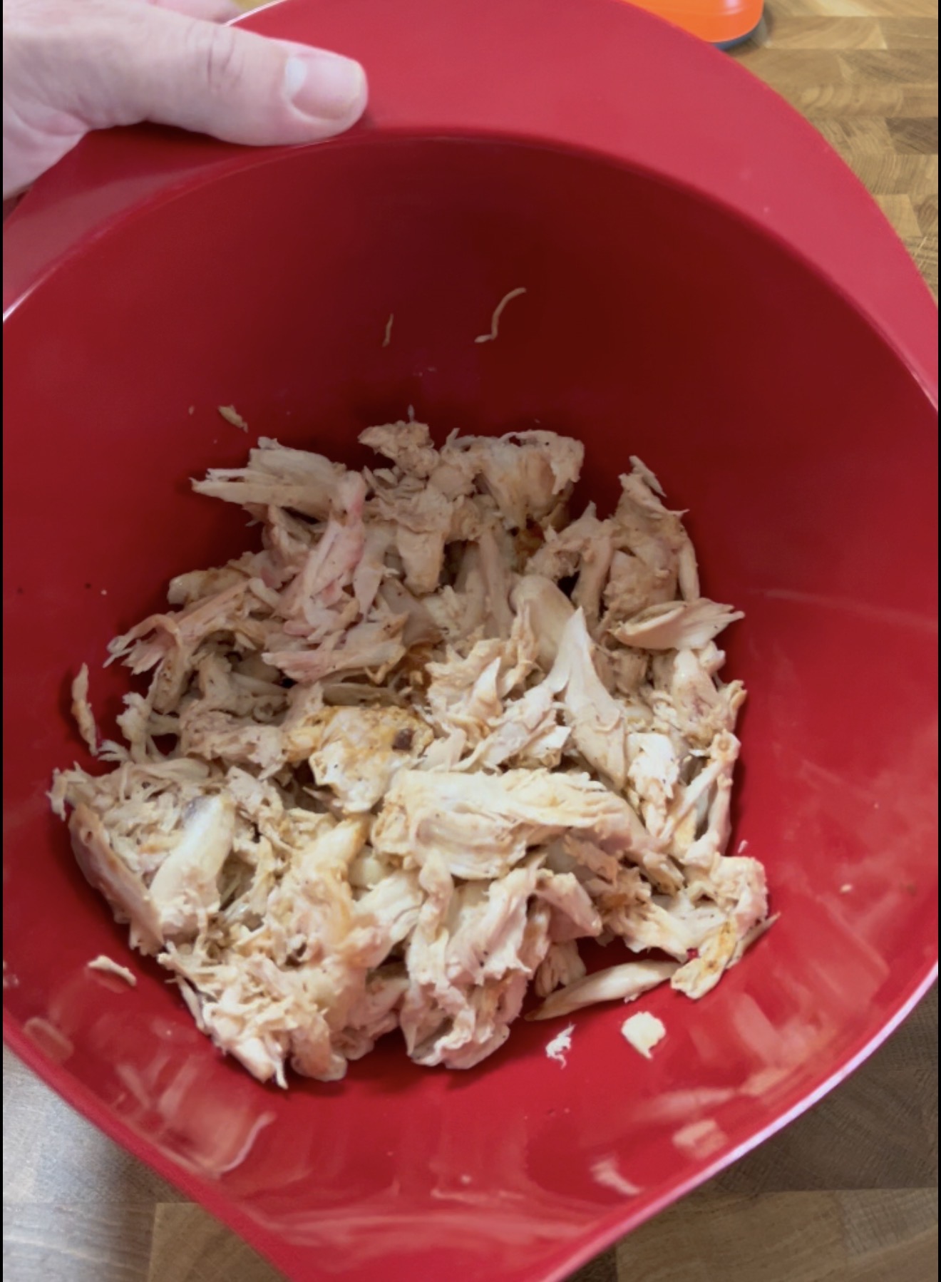 Shredded cooked chicken in a large red mixing bowl, held by a hand, on a wooden kitchen surface.