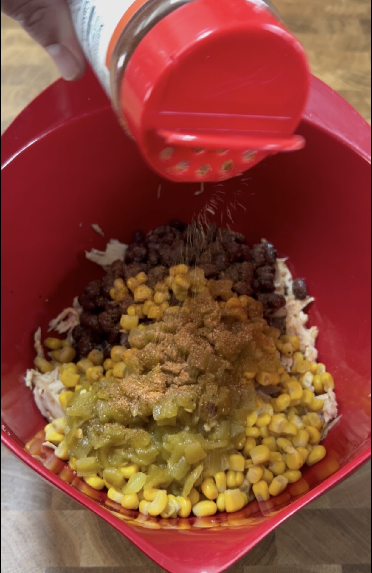 A red bowl contains shredded chicken, black beans, corn, and green chiles. A hand is sprinkling seasoning from a red spice container over the mixture.