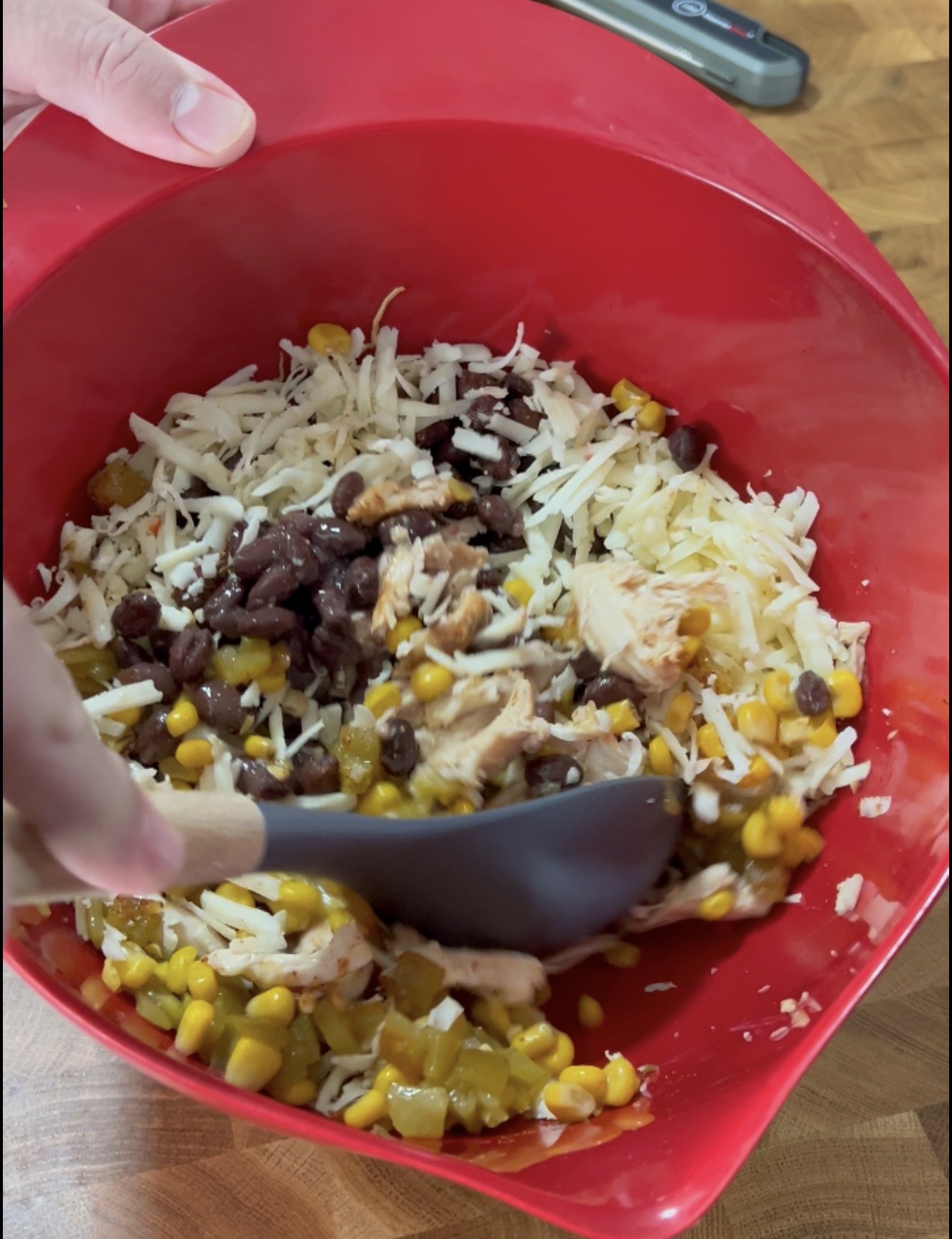 A hand stirs a mixture of shredded cheese, black beans, corn, and chicken in a red mixing bowl with a black spatula.