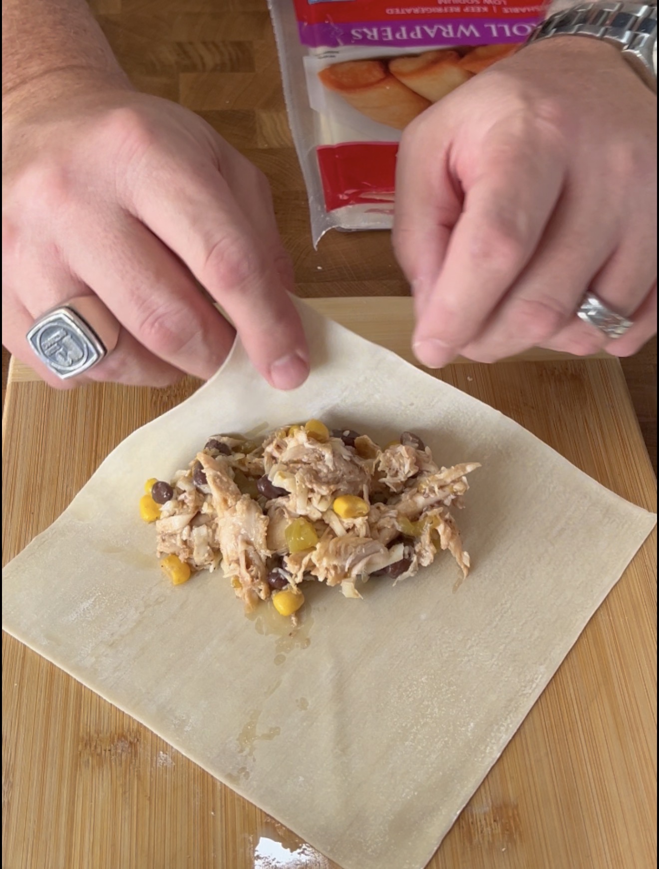 Hands folding an egg roll wrapper around a filling of shredded chicken, corn, and black beans on a wooden cutting board. An unopened package of egg roll wrappers is in the background.