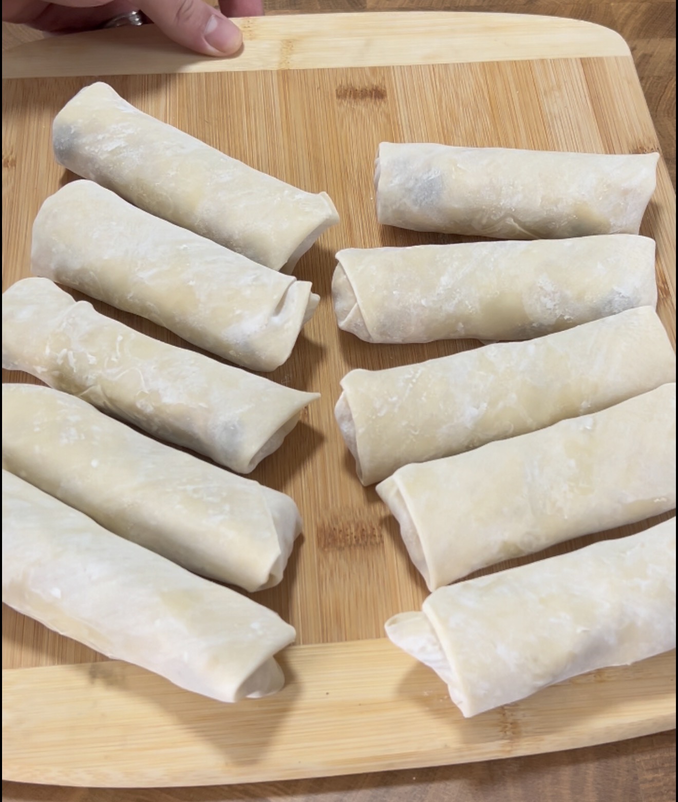 Nine uncooked spring rolls are arranged on a wooden cutting board. A hand is visible at the top left, holding the board. The spring rolls are wrapped in a light dough and dusted with flour.