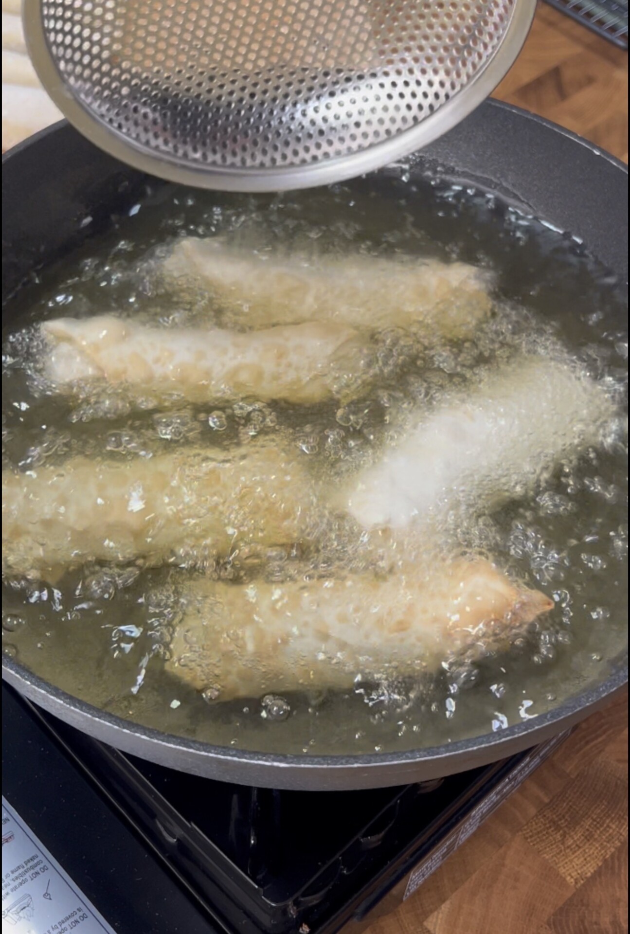 Four spring rolls are being deep-fried in hot oil in a pan, with a slotted lid partially covering the pan. The rolls are turning golden brown as they cook.