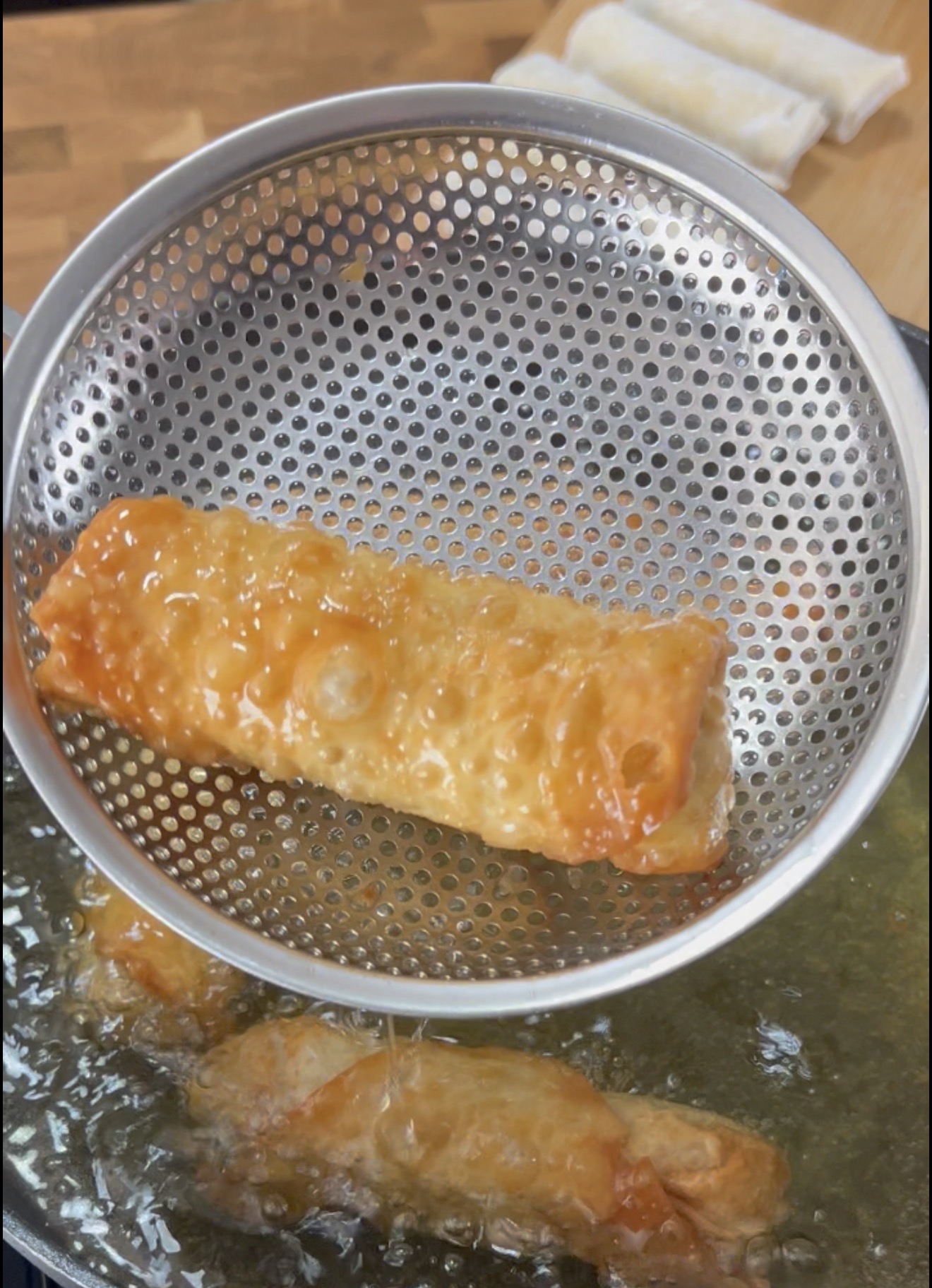 A freshly fried spring roll is held above hot oil in a metal strainer. Another roll fries in the oil below, and uncooked spring rolls are visible on a wooden surface in the background.