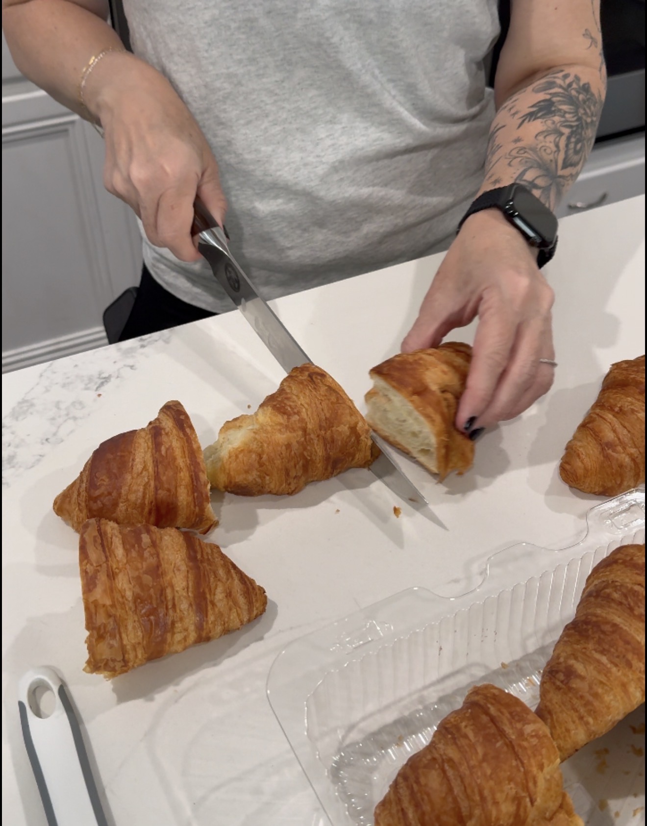 A person with tattoos and a smartwatch slices a croissant on a white countertop, with several other croissants and a plastic container nearby.