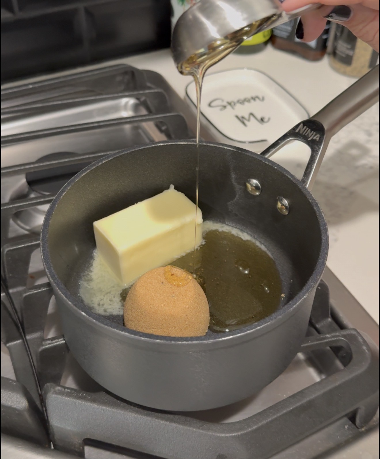 A saucepan on a stove contains a stick of butter and brown sugar. A hand pours syrup or honey from a measuring cup into the pan. A small dish labeled “Spoon Me” is in the background.