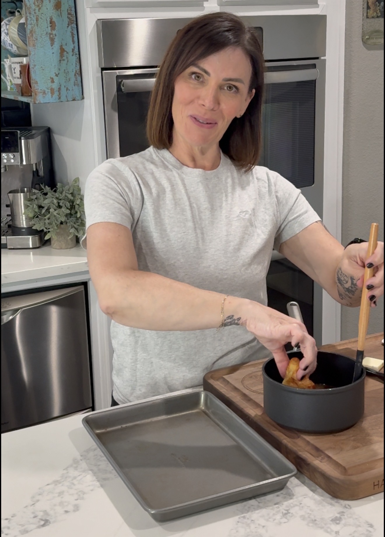 A woman stands in a kitchen, dipping food into a saucepan on a wooden cutting board. She holds a wooden utensil and smiles at the camera. A metal baking tray is on the counter in front of her.