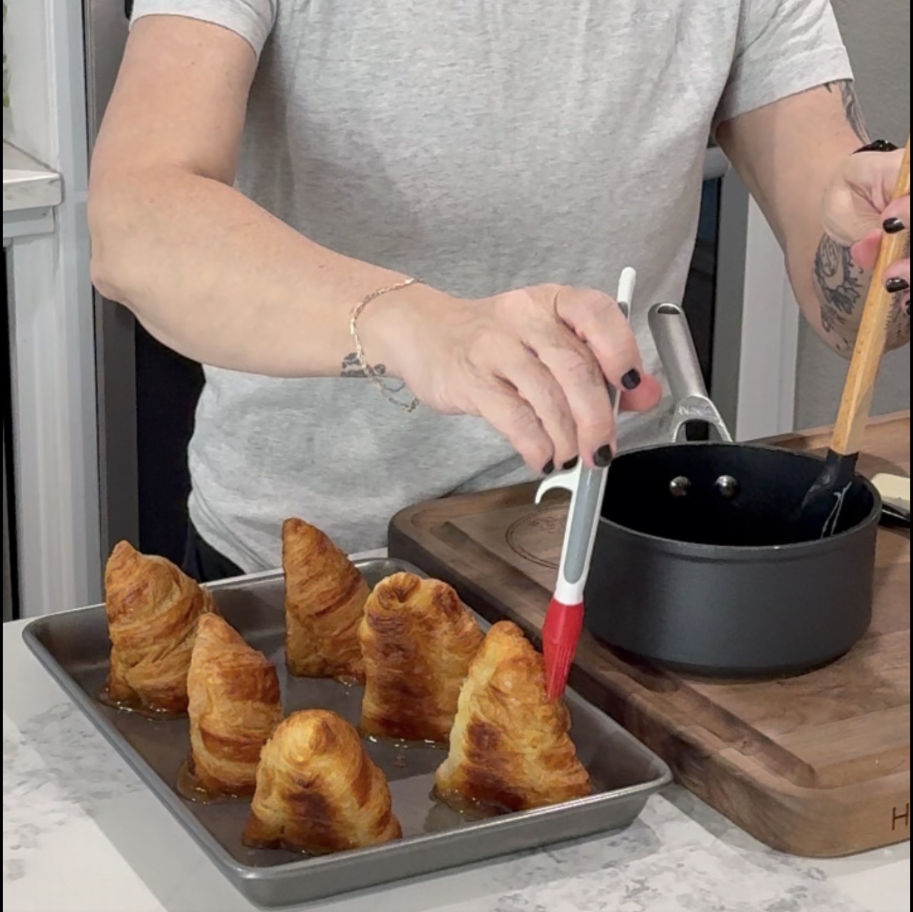A person brushes syrup onto six cone-shaped pastries on a baking tray, with a saucepan and spatula nearby on a wooden cutting board.