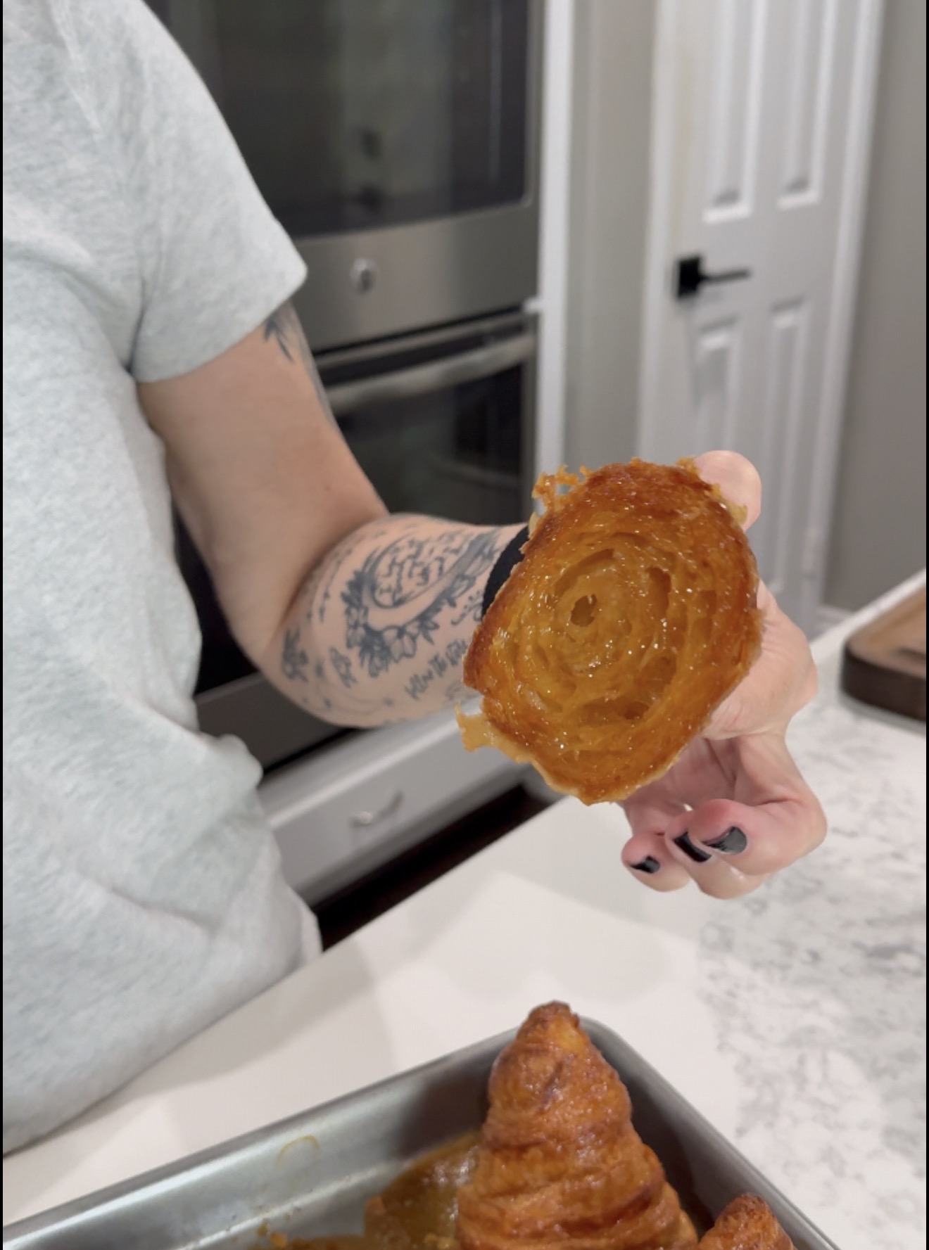 A person with tattoos holds a glazed, spiral pastry in a kitchen, with an oven and white cabinets in the background.