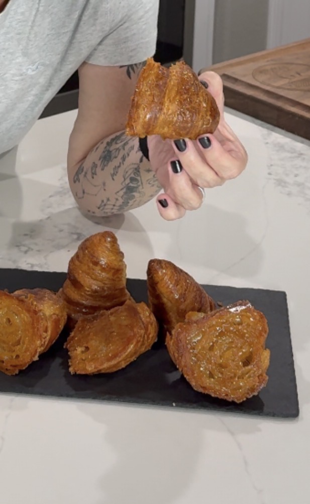 A person with black nail polish and a tattooed arm holds a pastry above a slate tray with four golden, flaky pastries on a white countertop.