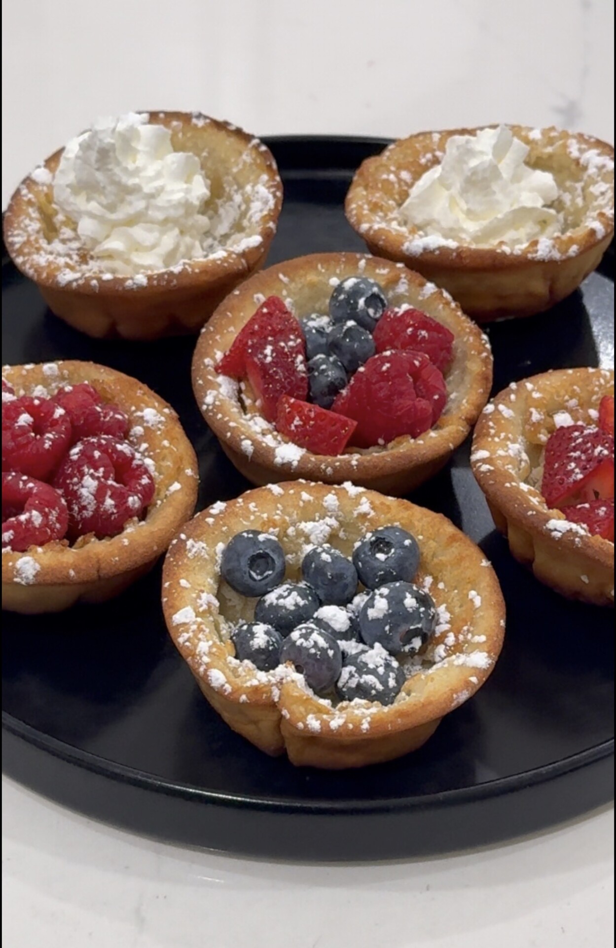 Six mini tartlets on a black plate, topped with whipped cream, blueberries, raspberries, and strawberries, all dusted with powdered sugar.