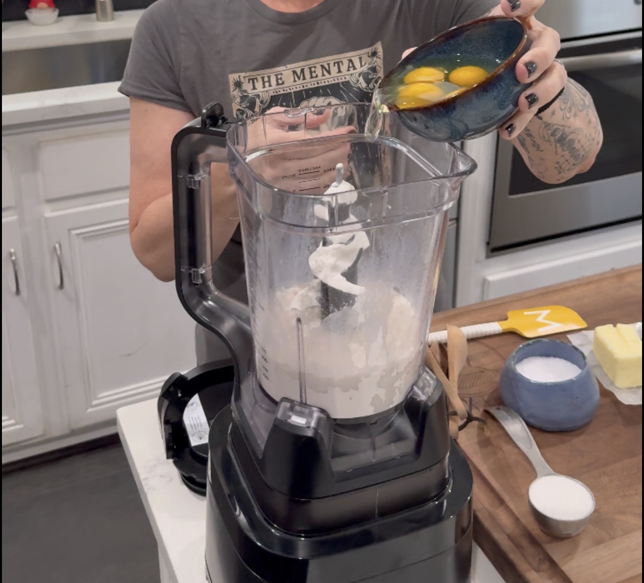 A person pours eggs from a blue bowl into a food processor containing flour. Other ingredients and utensils are on a wooden counter nearby.