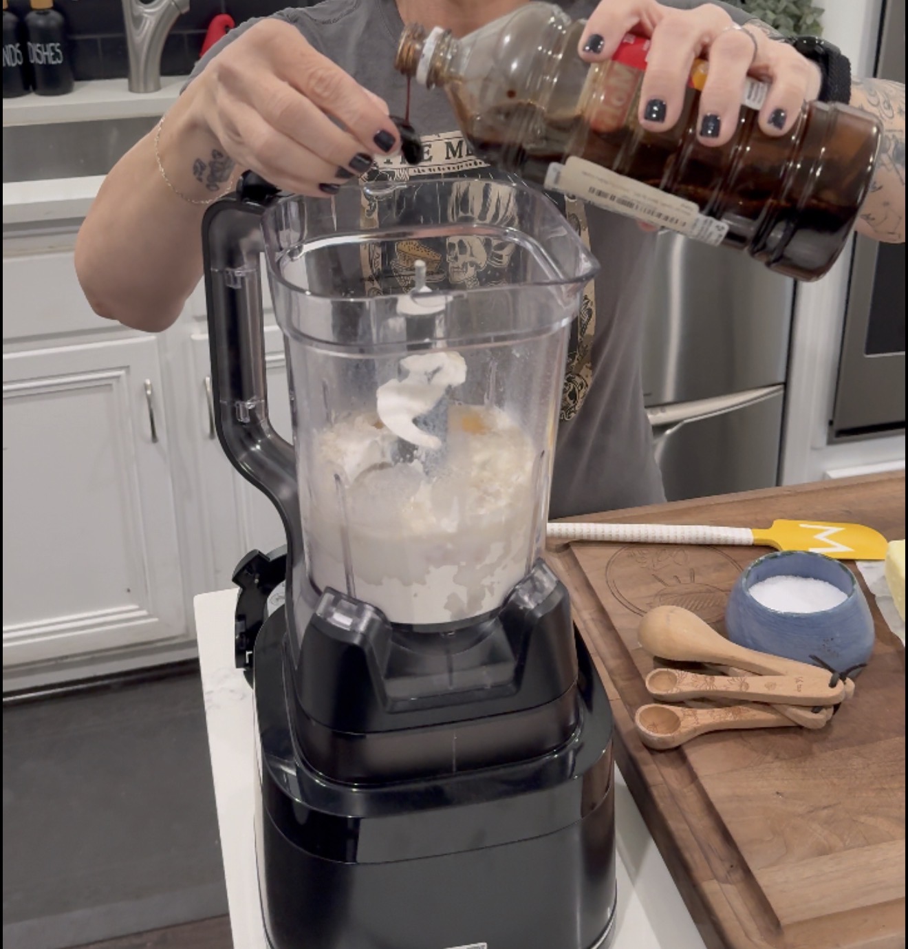 A person pours vanilla extract into a food processor containing flour and other ingredients. Measuring spoons and a spatula are on a wooden cutting board nearby.