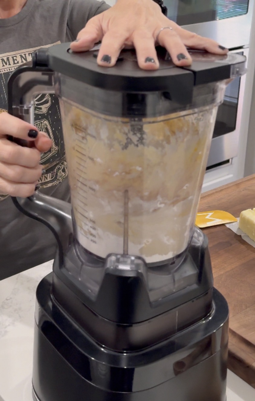 A person holds the lid of a blender filled with ingredients as it mixes, preparing food in a kitchen. Only their hands and part of their shirt are visible.