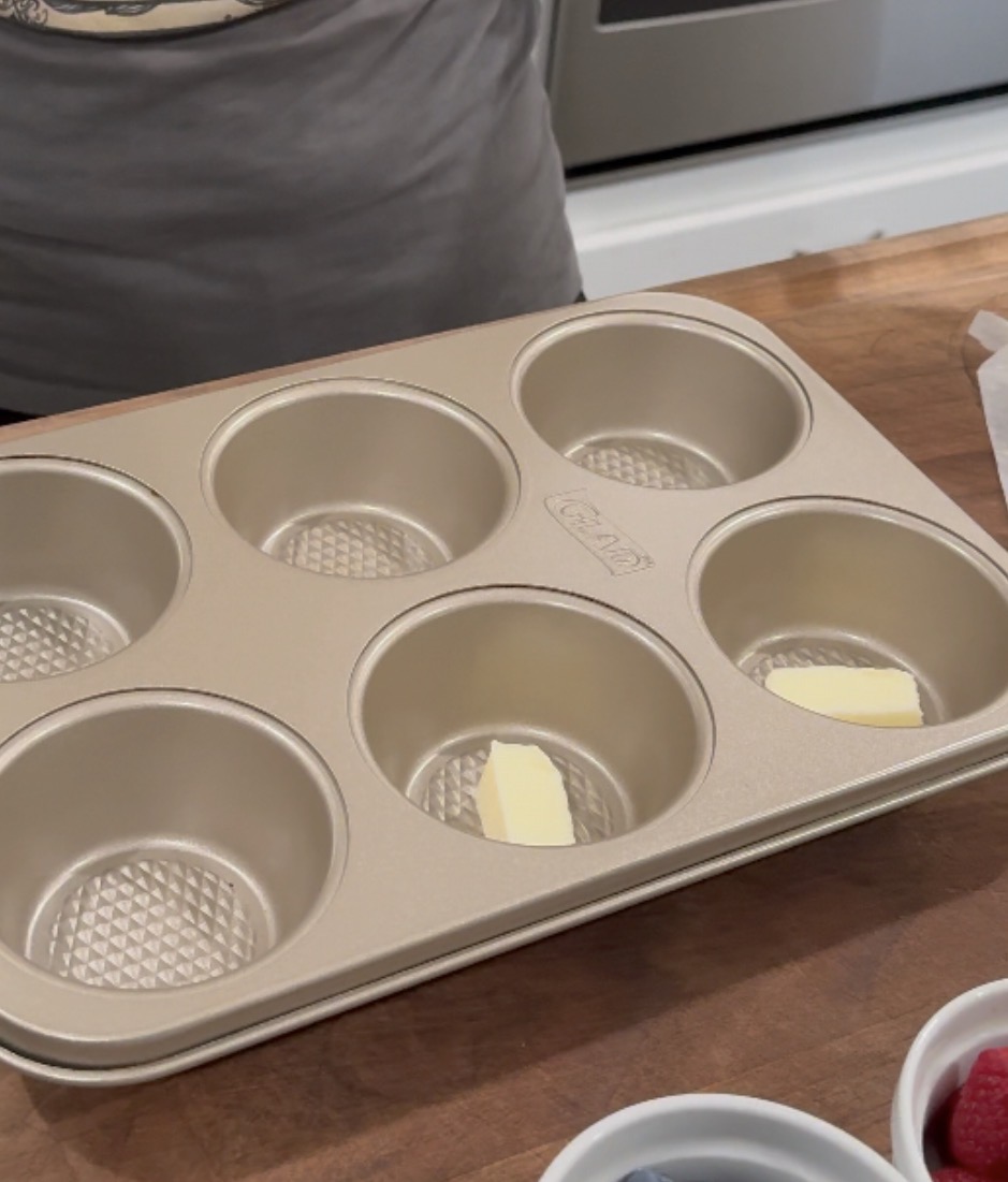 A six-cup muffin pan with two cups holding small pieces of butter sits on a wooden countertop near a bowl of berries.