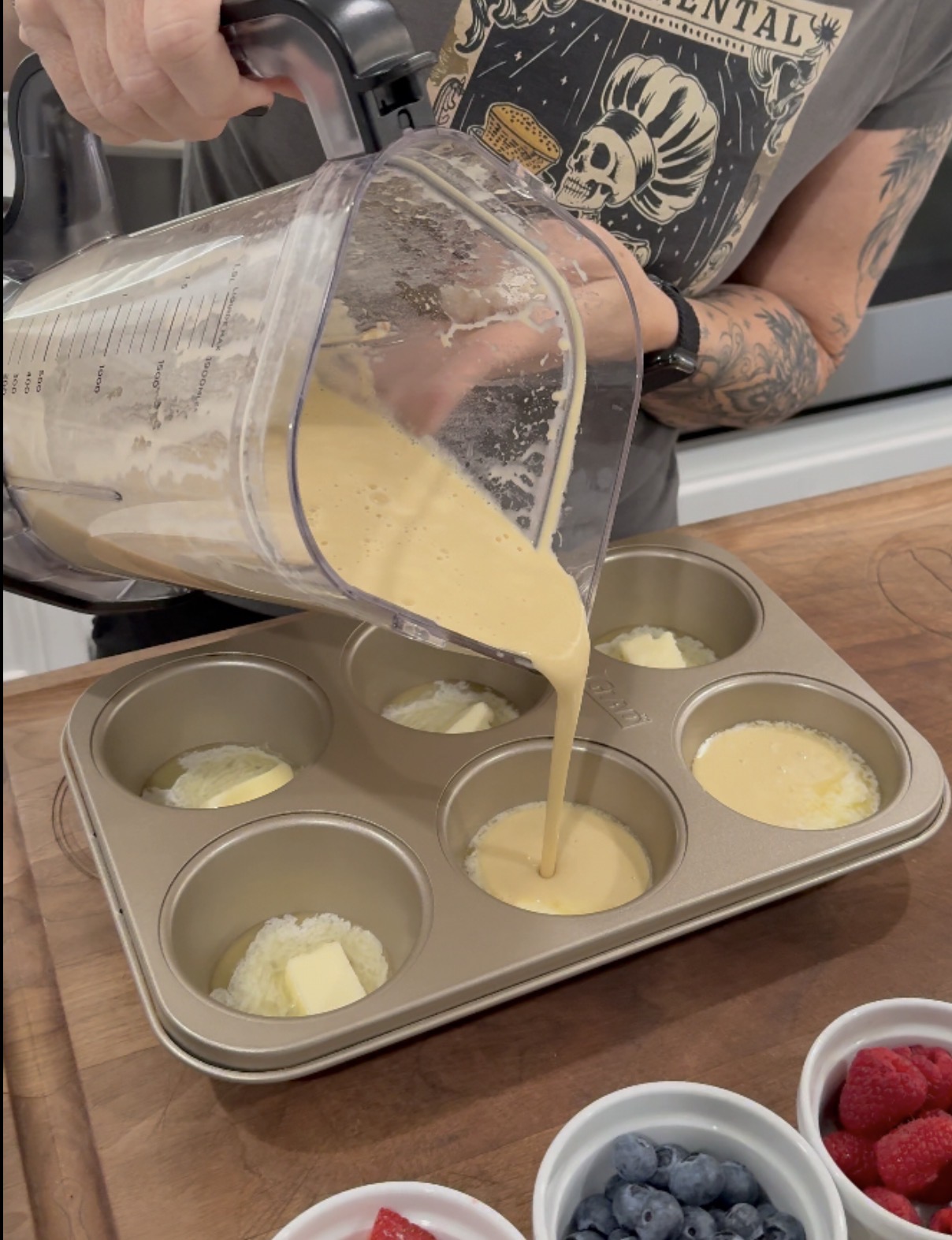 A person pours batter from a blender into a muffin tin on a wooden counter, with bowls of butter, blueberries, raspberries, and strawberries nearby.