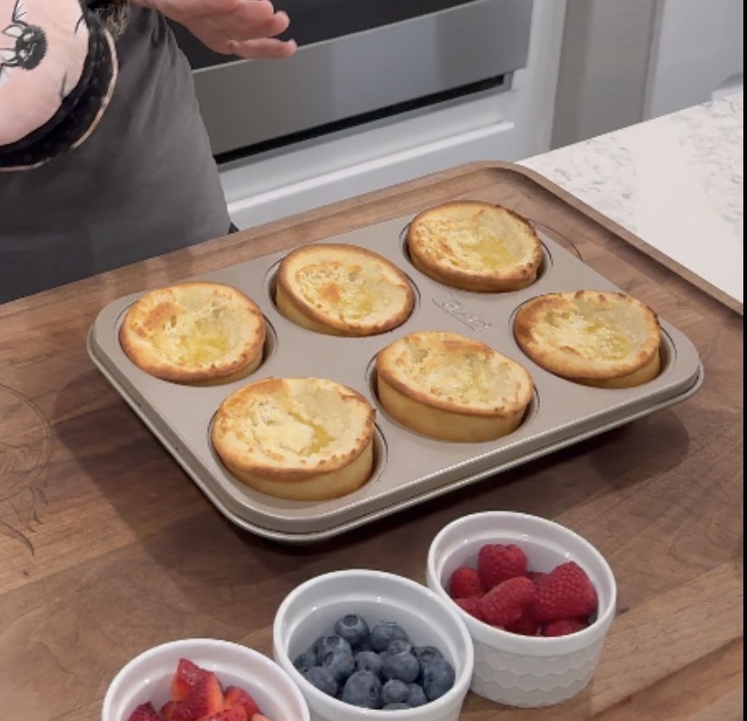 A muffin tin with six baked pancakes sits on a wooden counter. Nearby are small bowls of sliced strawberries, blueberries, and raspberries. A person’s hand is visible above the tin.