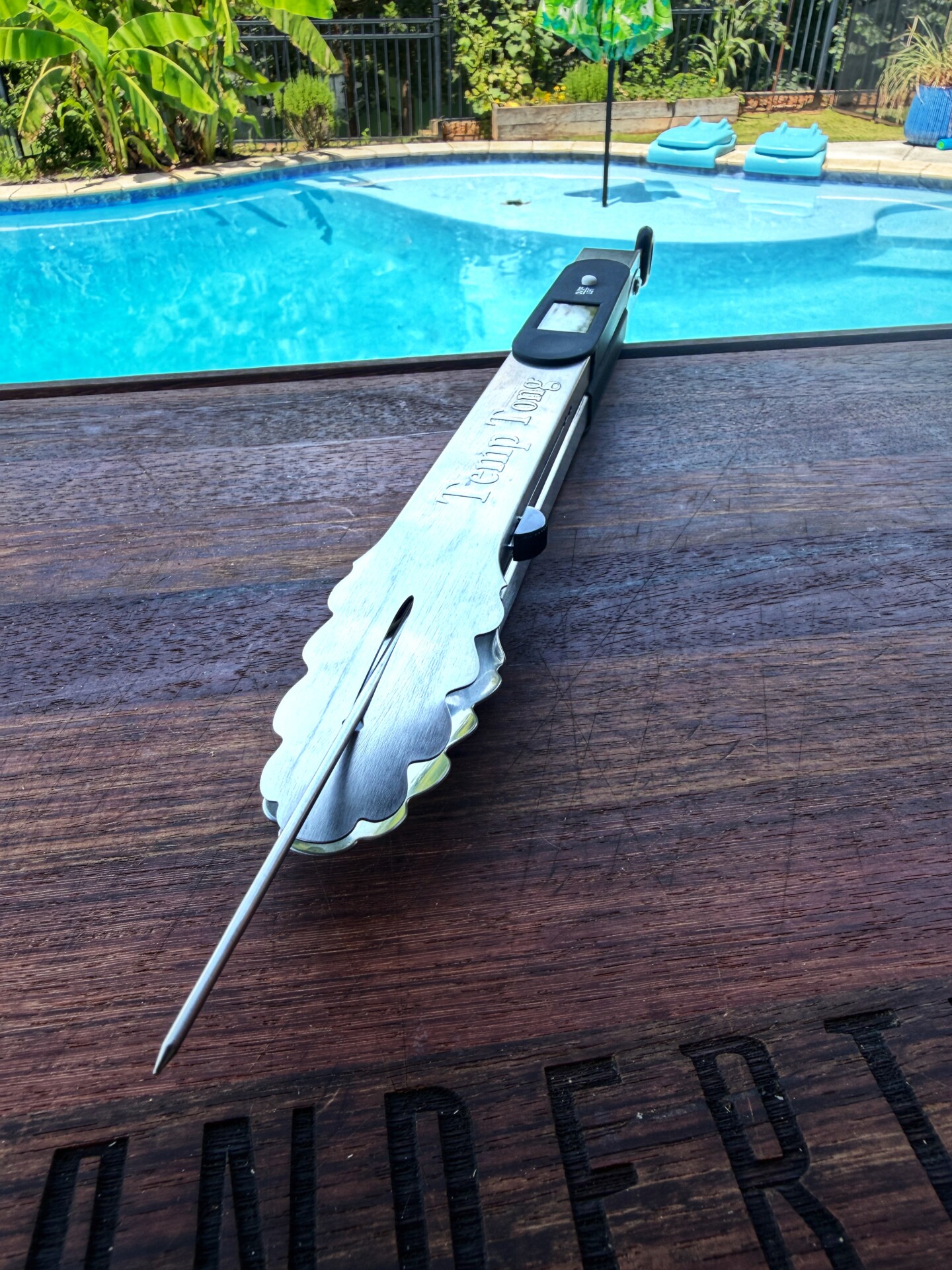A pair of metal BBQ tongs with a built-in meat thermometer rests on a wooden table beside a swimming pool, with greenery and pool floats in the background.