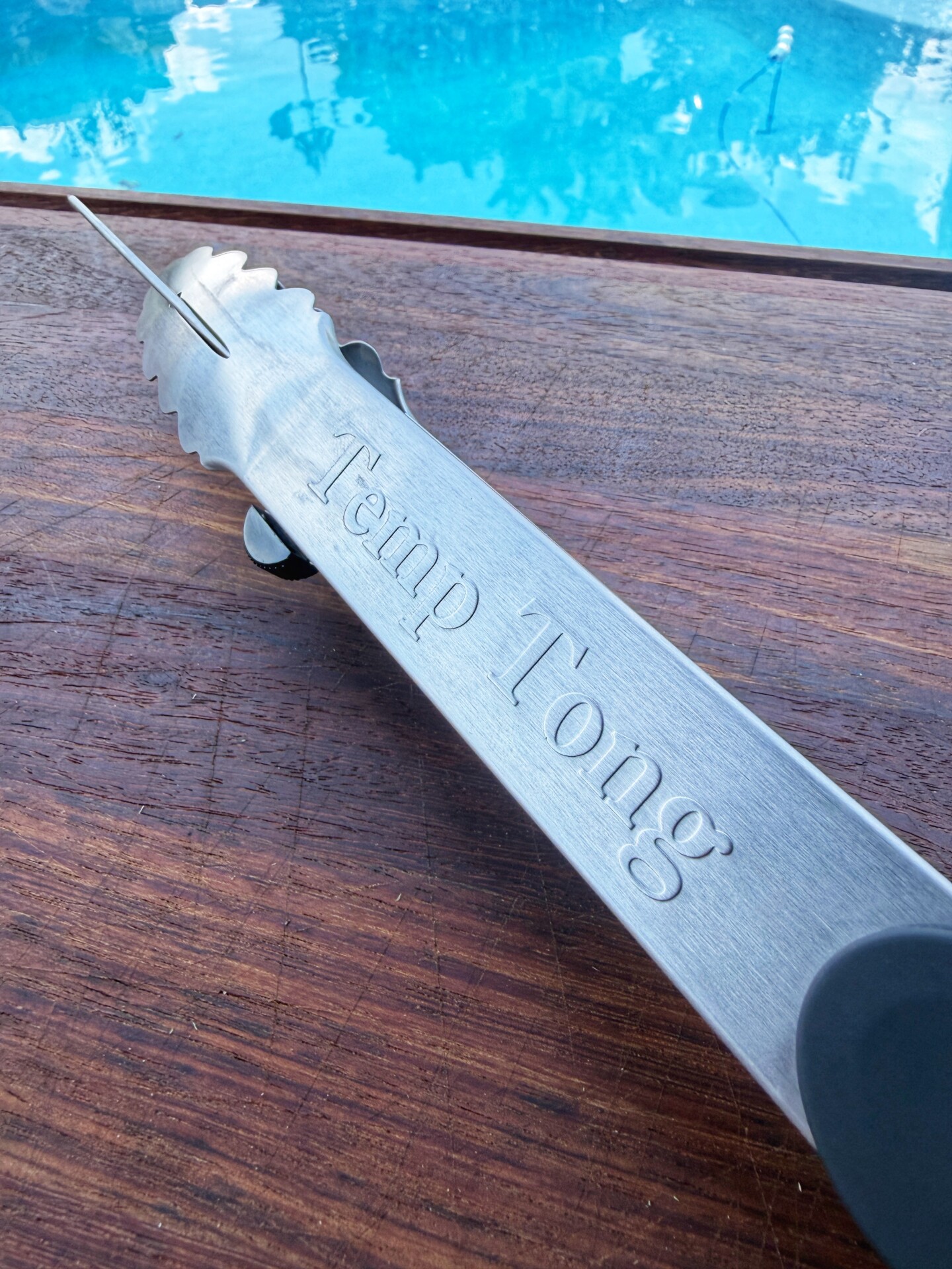 A pair of stainless steel tongs labeled Temp Tong rests on a wooden surface near a pool, with a built-in meat thermometer visible at the gripping end.