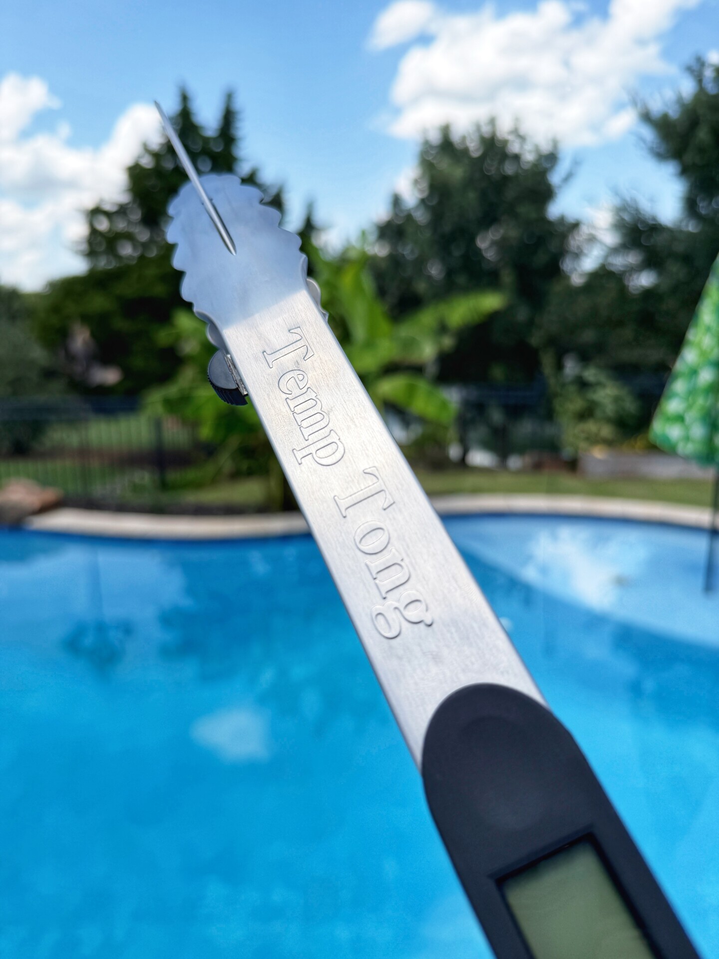 A pair of metal tongs labeled Temp Tong held above a blue swimming pool, with trees, a fence, and a cloudy sky in the background.