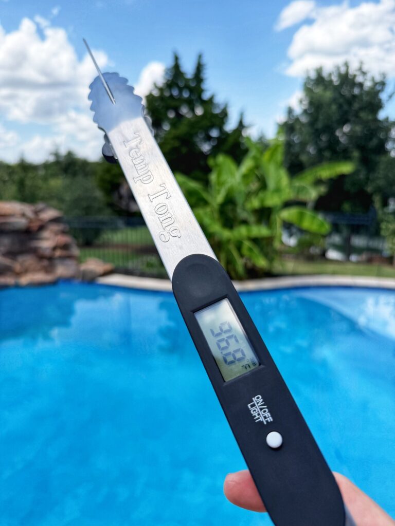 A hand holds metal grill tongs with a digital thermometer reading 36.5°C by a blue swimming pool, with trees and a partly cloudy sky in the background.