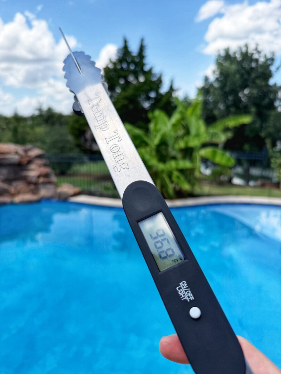 A hand holds metal grill tongs with a digital thermometer reading 36.5°C by a blue swimming pool, with trees and a partly cloudy sky in the background.