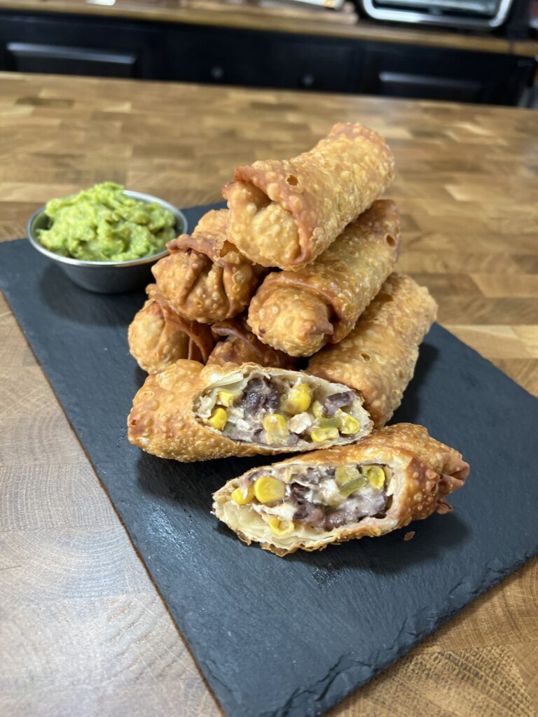 A stack of crispy egg rolls filled with black beans, corn, and cheese sits on a slate board, with a small bowl of guacamole on the side.