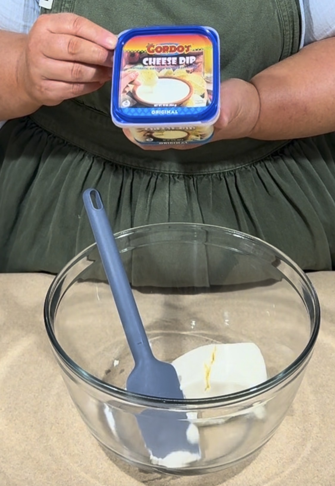 A person holds a container of Cordos Cheese Dip above a glass mixing bowl with a blue spatula and a block of cream cheese inside.
