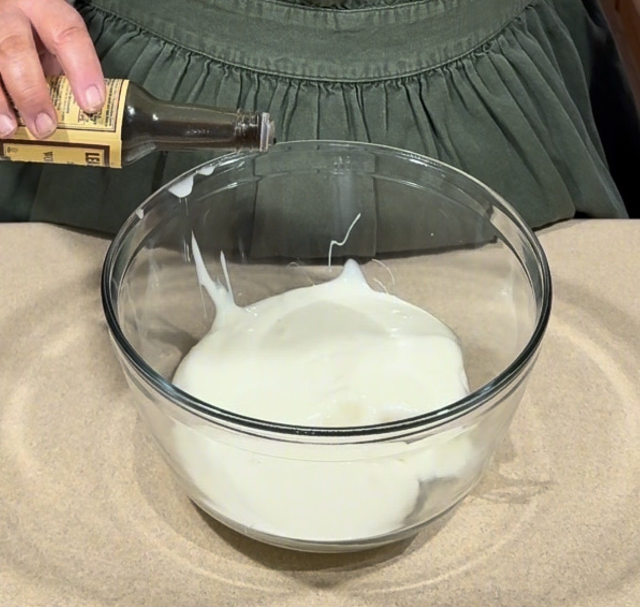 A person in a green apron pours vanilla extract from a small bottle into a glass bowl filled with a thick white mixture on a tan surface.