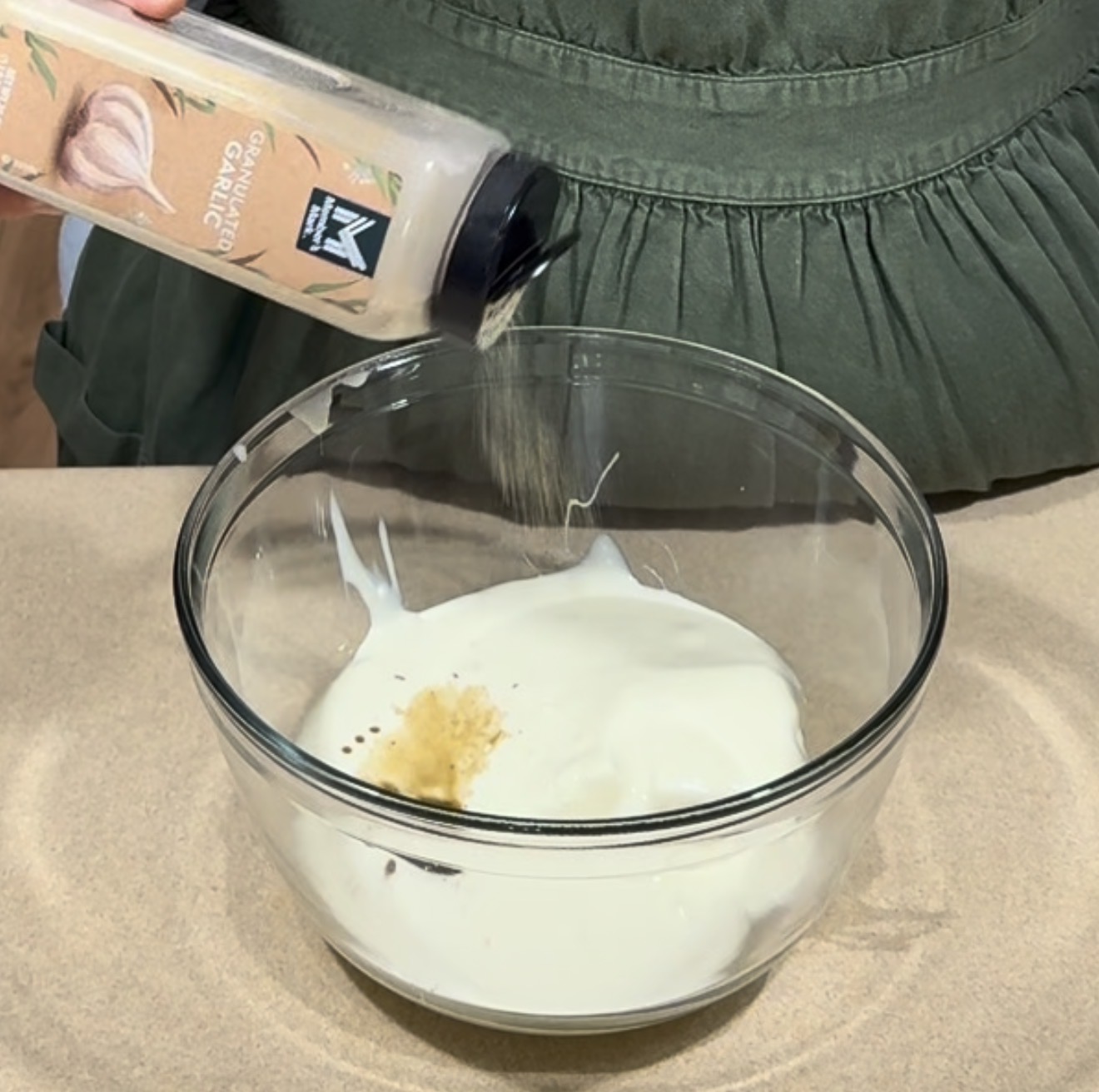 A person sprinkles garlic powder from a container into a glass bowl filled with a creamy white mixture, on a beige surface.