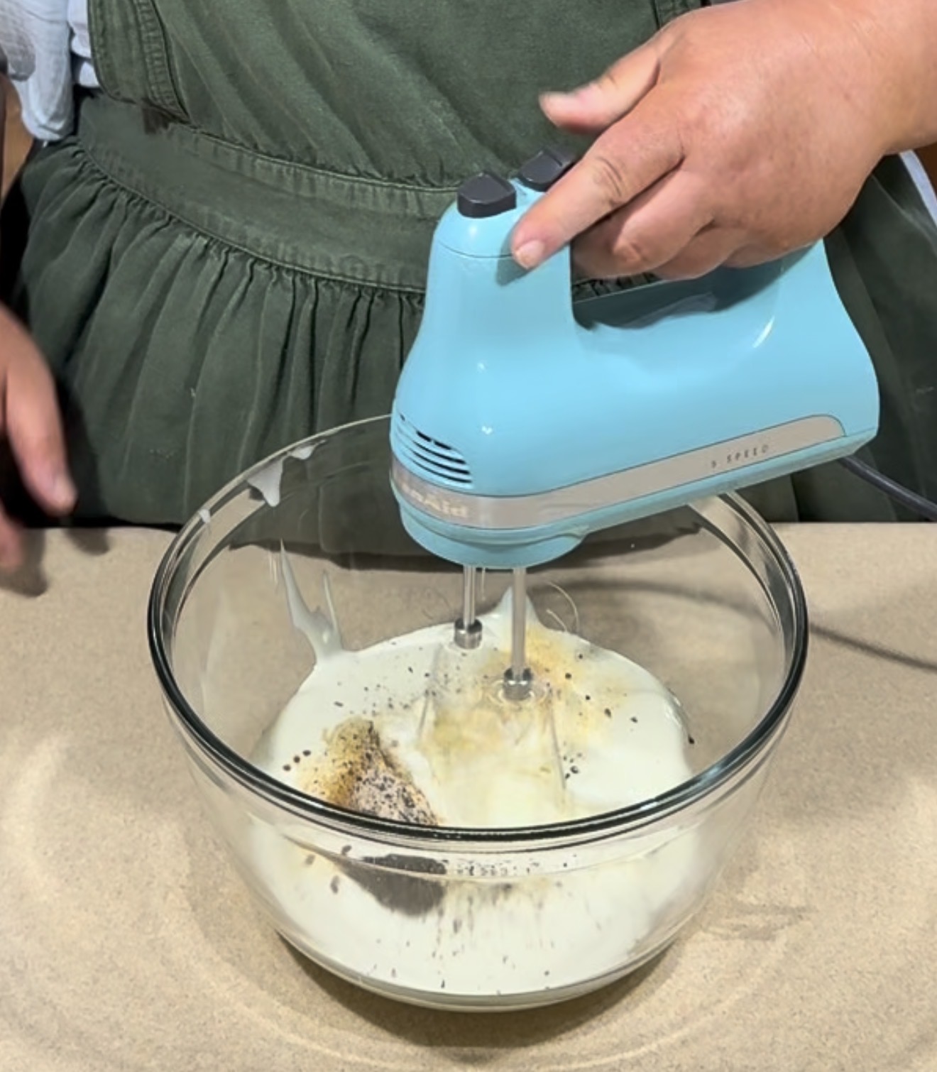 A person in a green apron uses a blue electric hand mixer to blend ingredients, including cream and spices, in a glass bowl on a beige countertop.