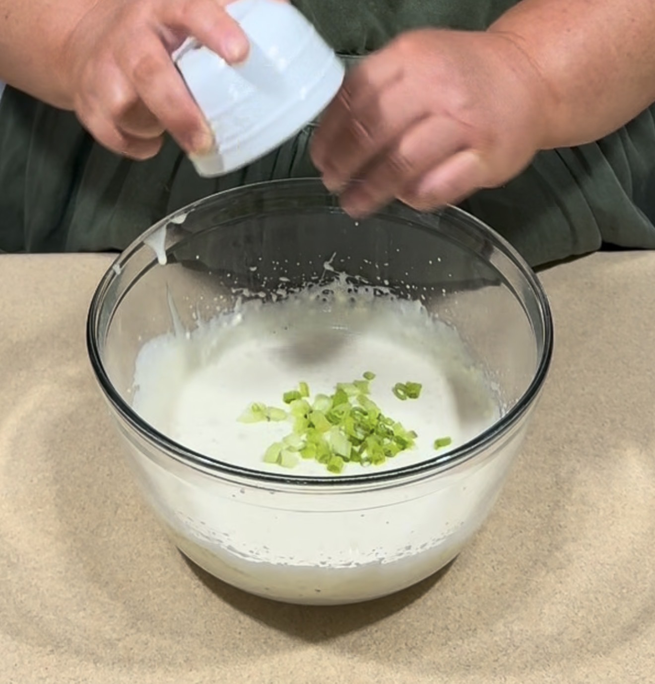 A person adds chopped green onions from a small bowl into a larger glass bowl filled with a creamy white mixture on a countertop.