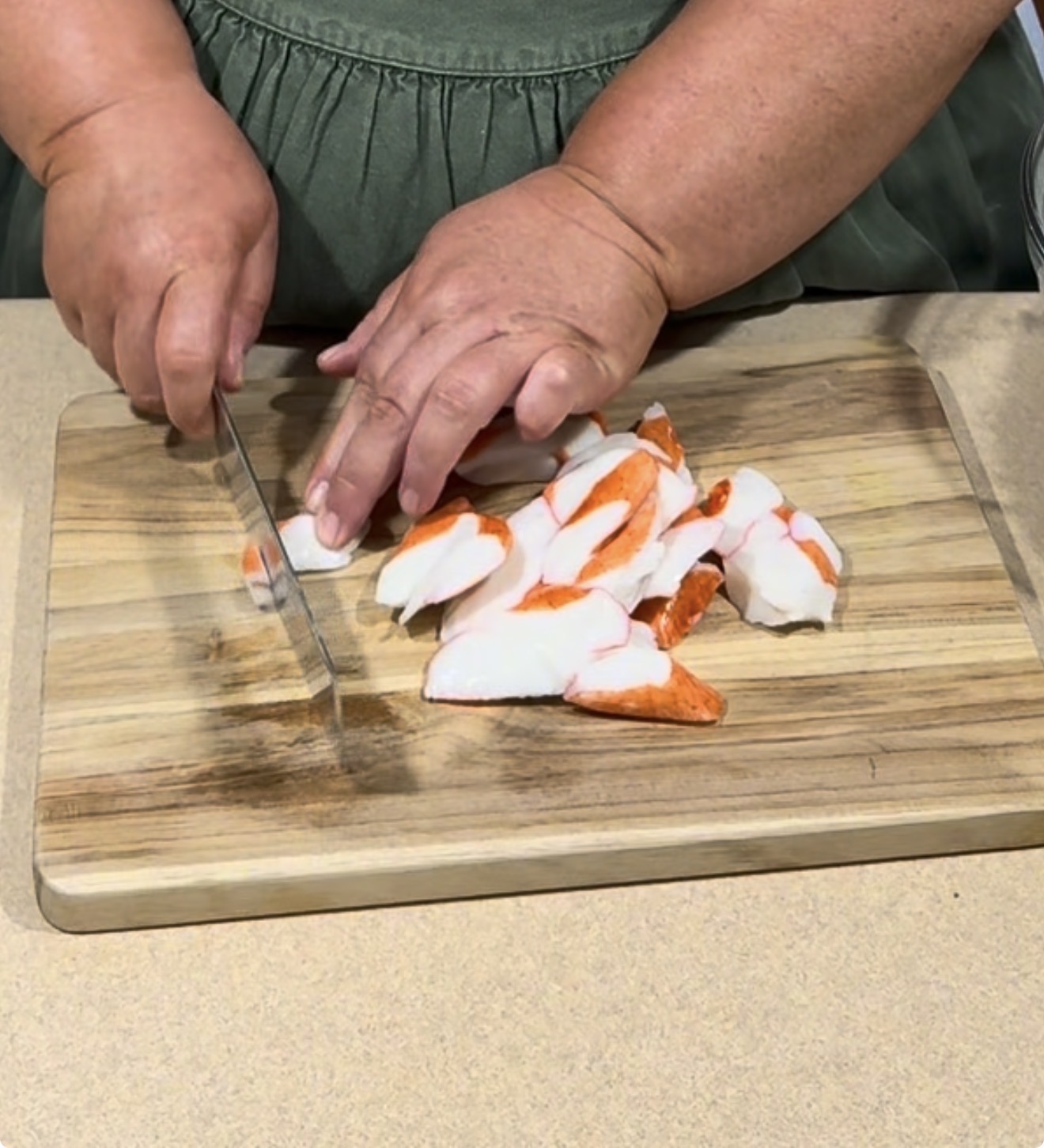 A person in a green apron slices imitation crab sticks on a wooden cutting board with a knife.