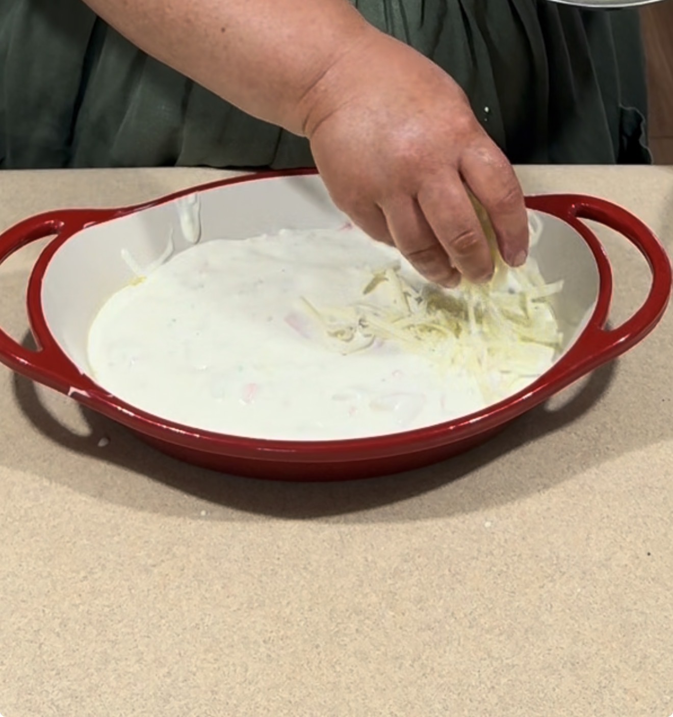 A person sprinkles shredded cheese over a creamy mixture in a red baking dish with handles, placed on a beige countertop.